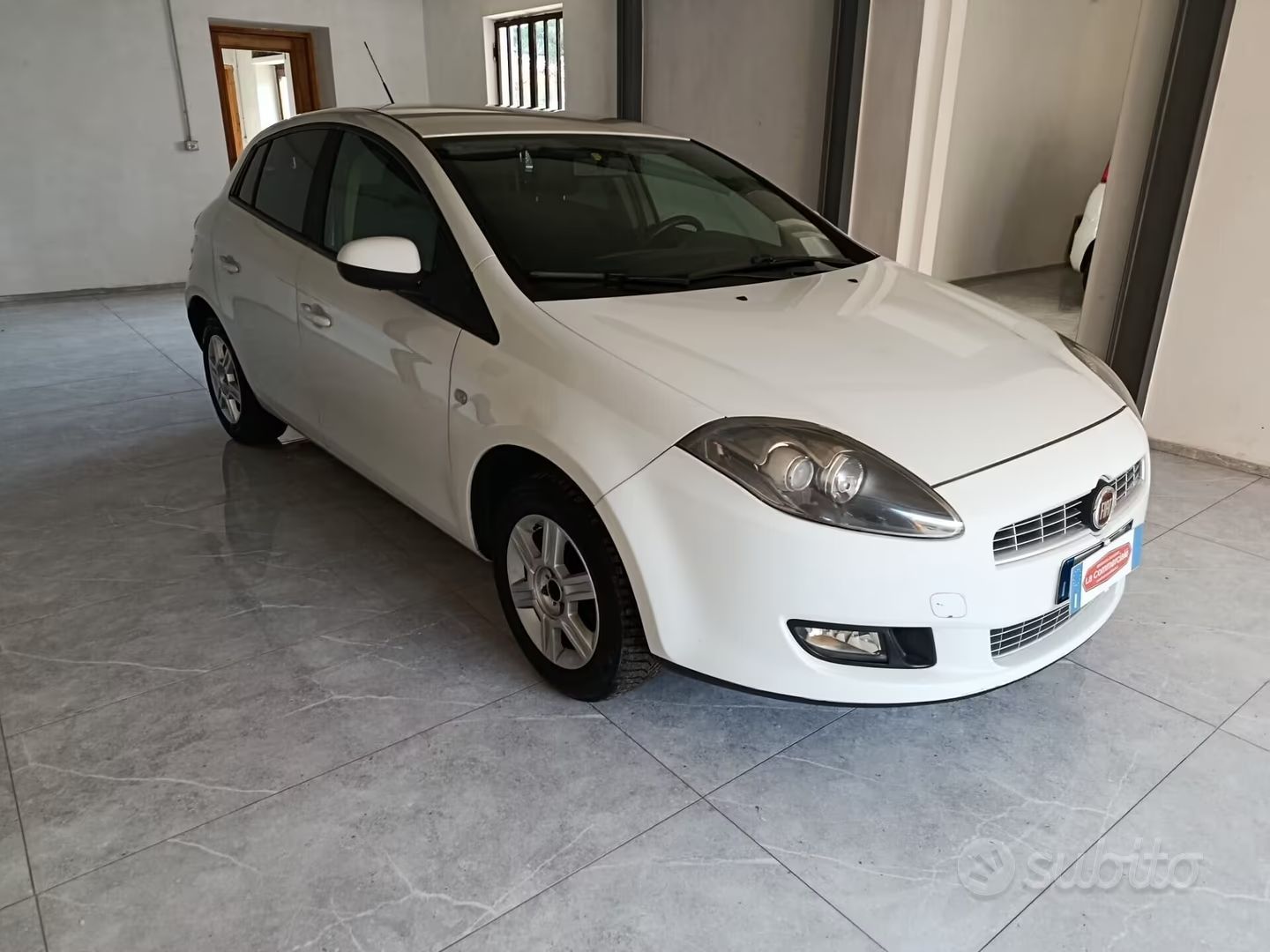 Auto hatchback bianca parcheggiata in un garage coperto con pavimento piastrellato.