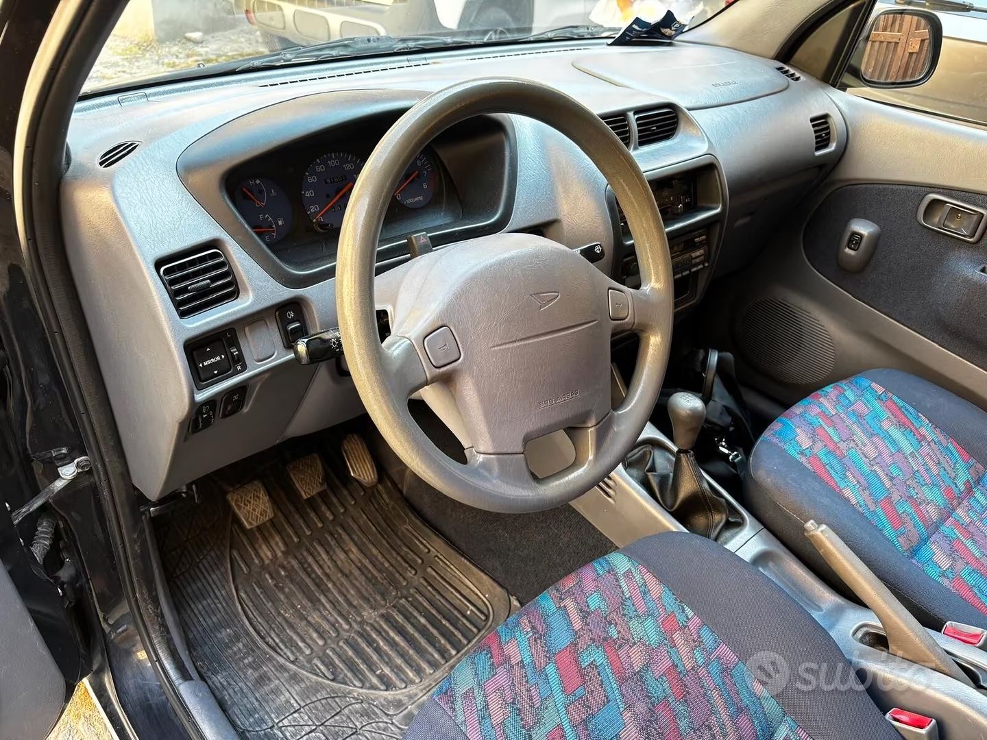 Interni dell'auto con volante, cruscotto, sedili con fantasia e tappetini usurati.