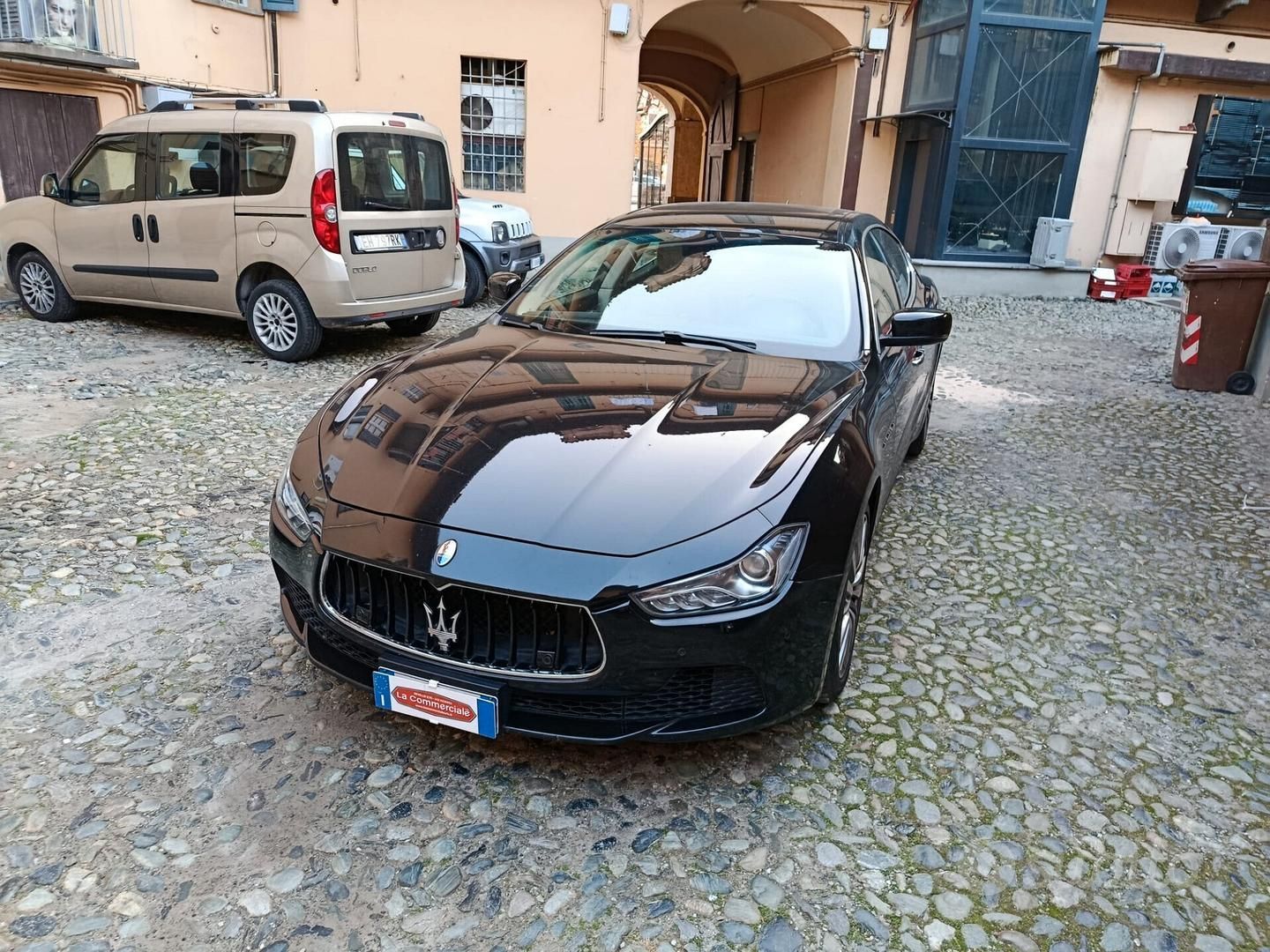 Una berlina sportiva nera parcheggiata in un cortile lastricato, accanto a edifici beige e un furgone.