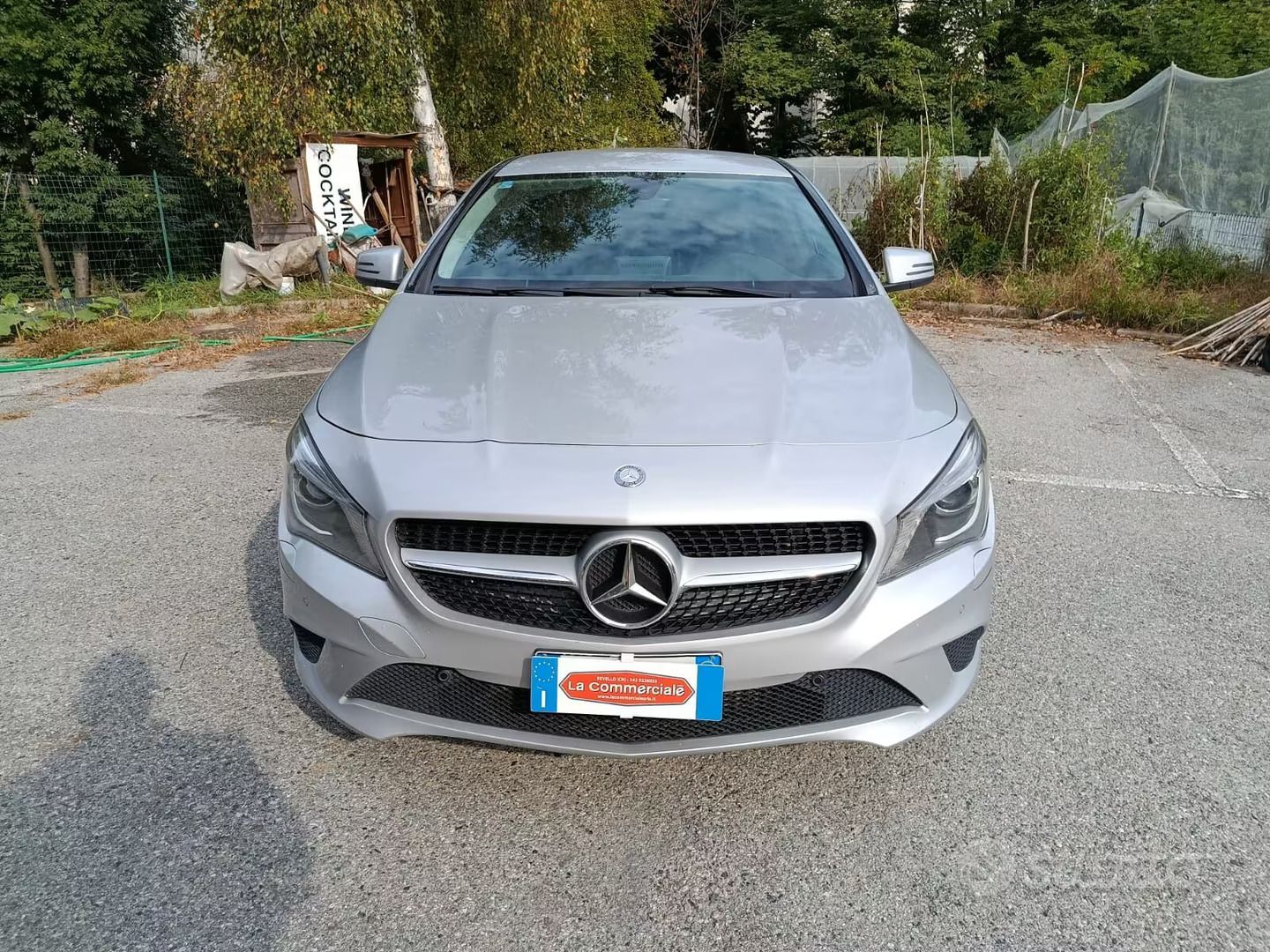 Vista frontale di una berlina Mercedes color argento parcheggiata su un vialetto di ghiaia, con alberi e recinzione sullo sfondo.