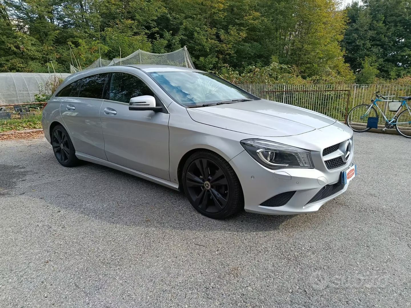 Berlina argentata parcheggiata su ghiaia vicino ad alberi e a una recinzione ricoperta di graffiti.