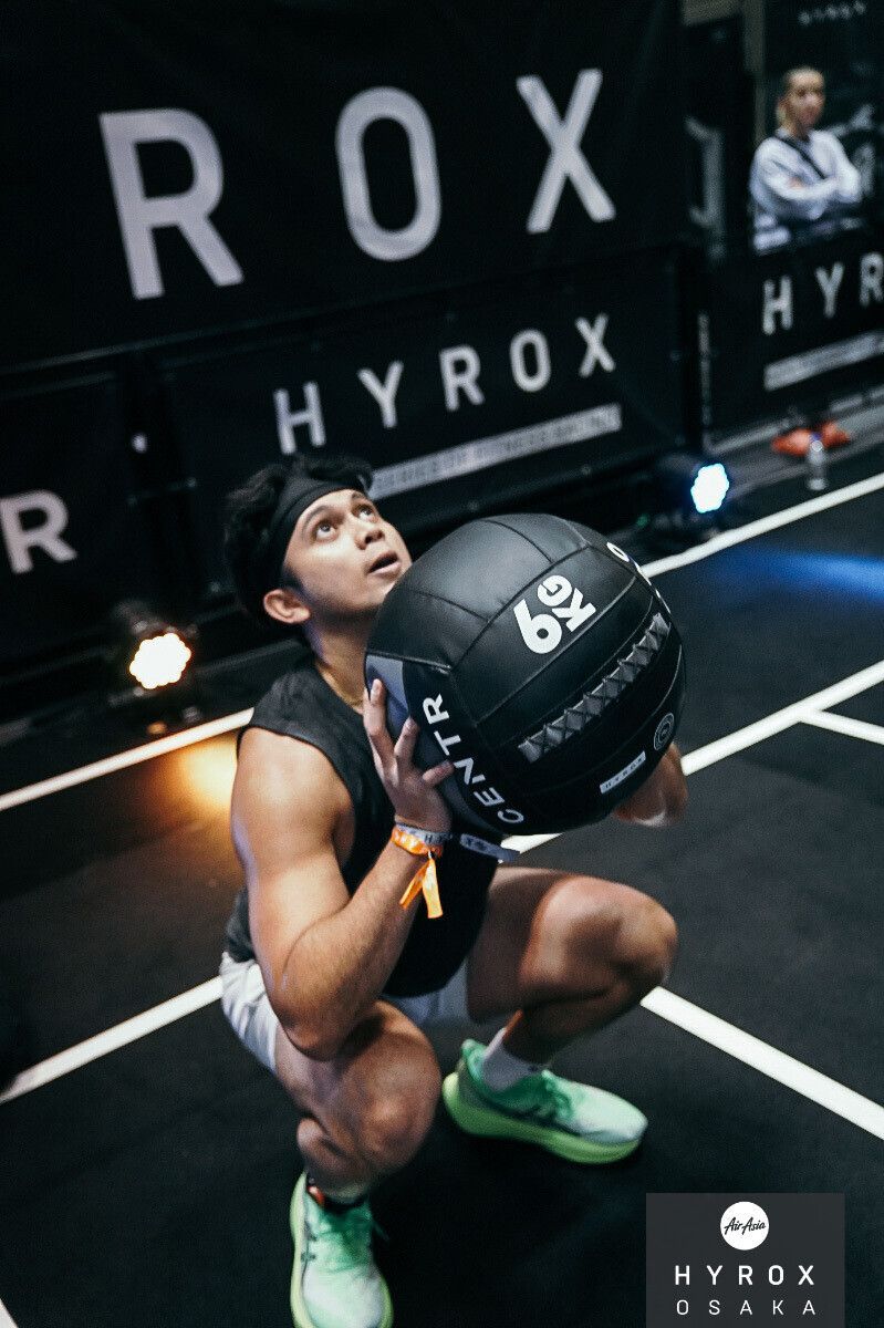 An athlete in a squat position holds a black medicine ball at a Hyrox fitness competition.