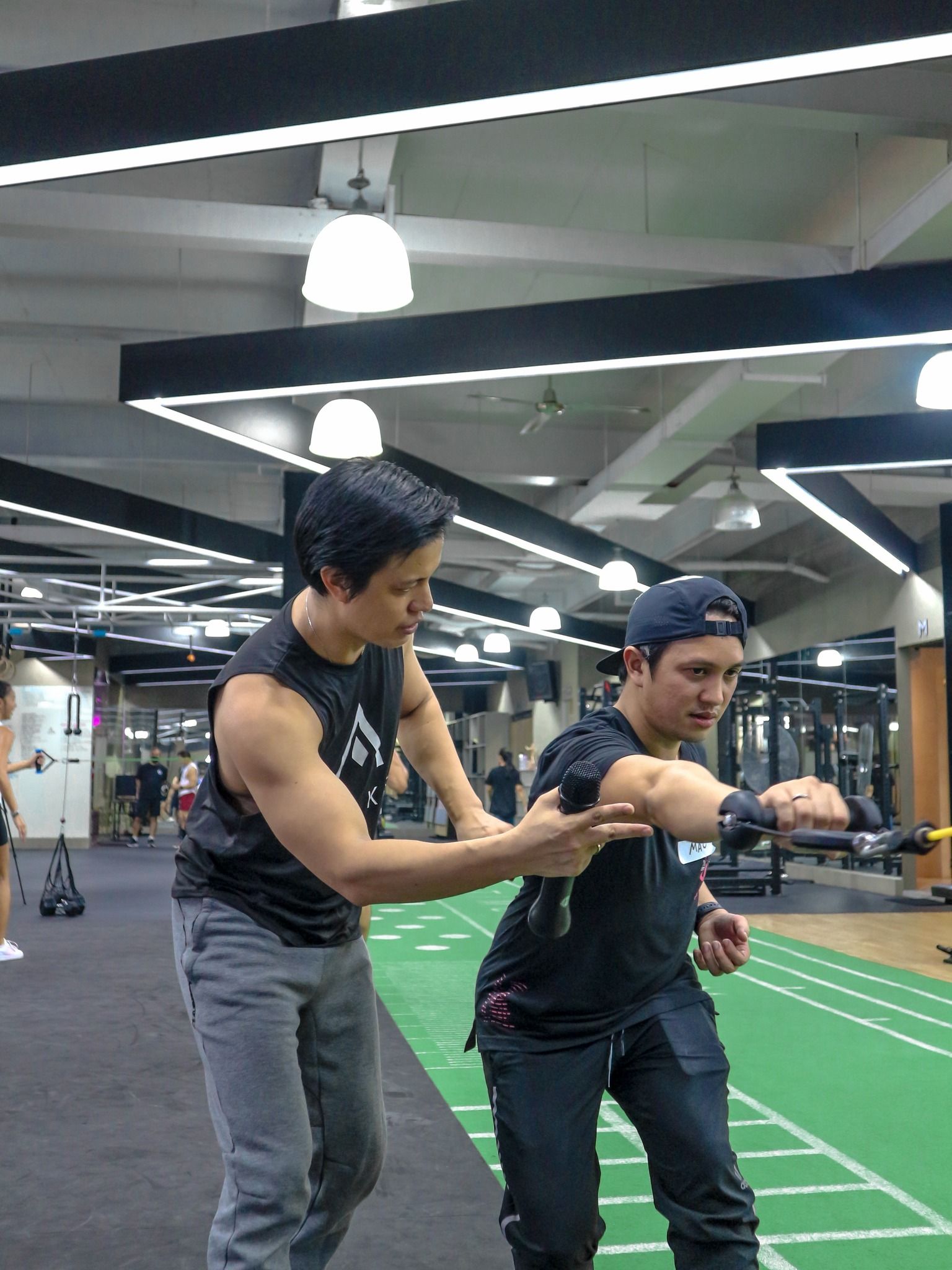 A trainer guides a person performing an arm exercise with a resistance band in a gym with overhead lighting.