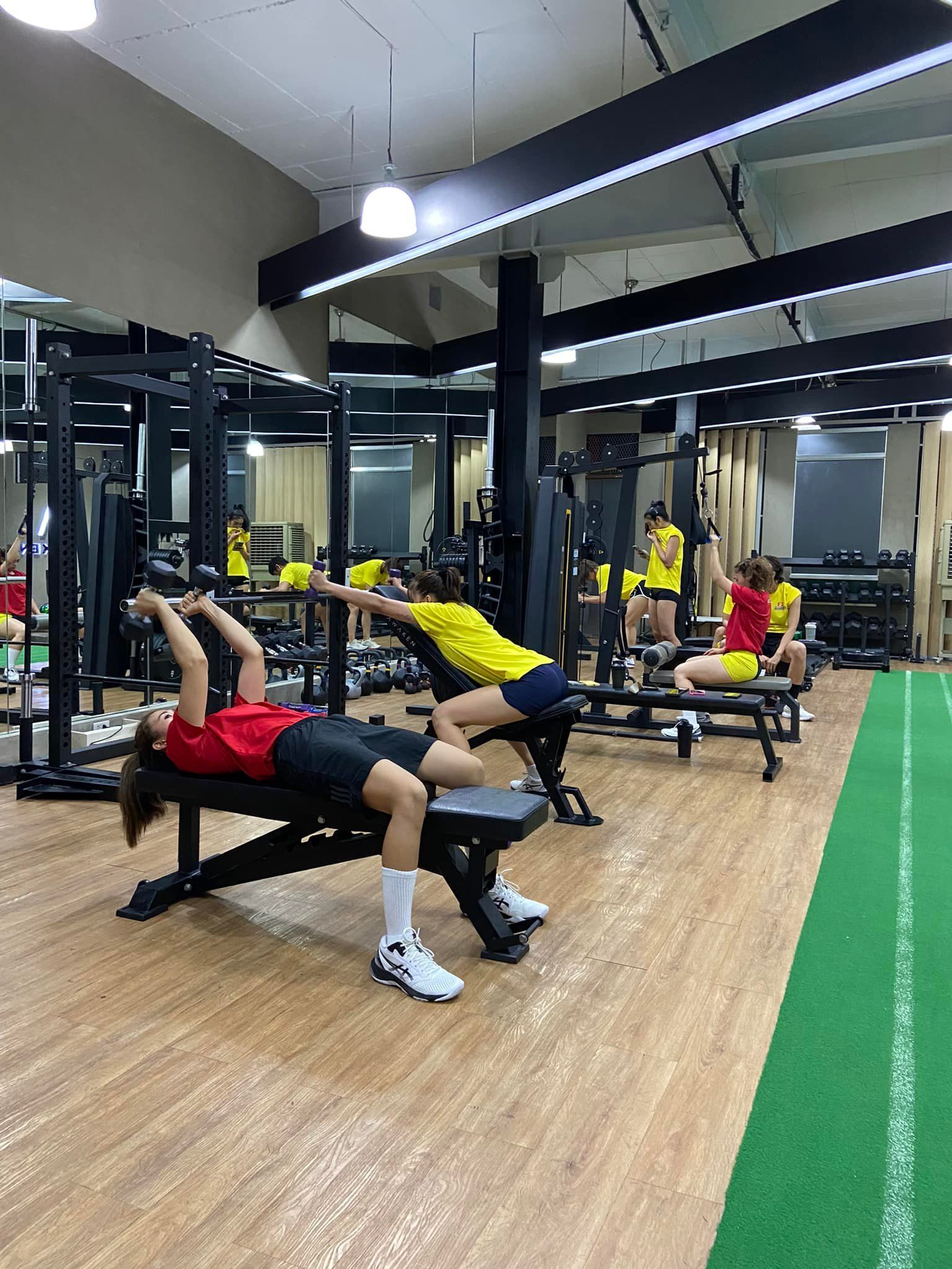 Athletes in a gym using benches to perform weightlifting exercises on a green turf floor.