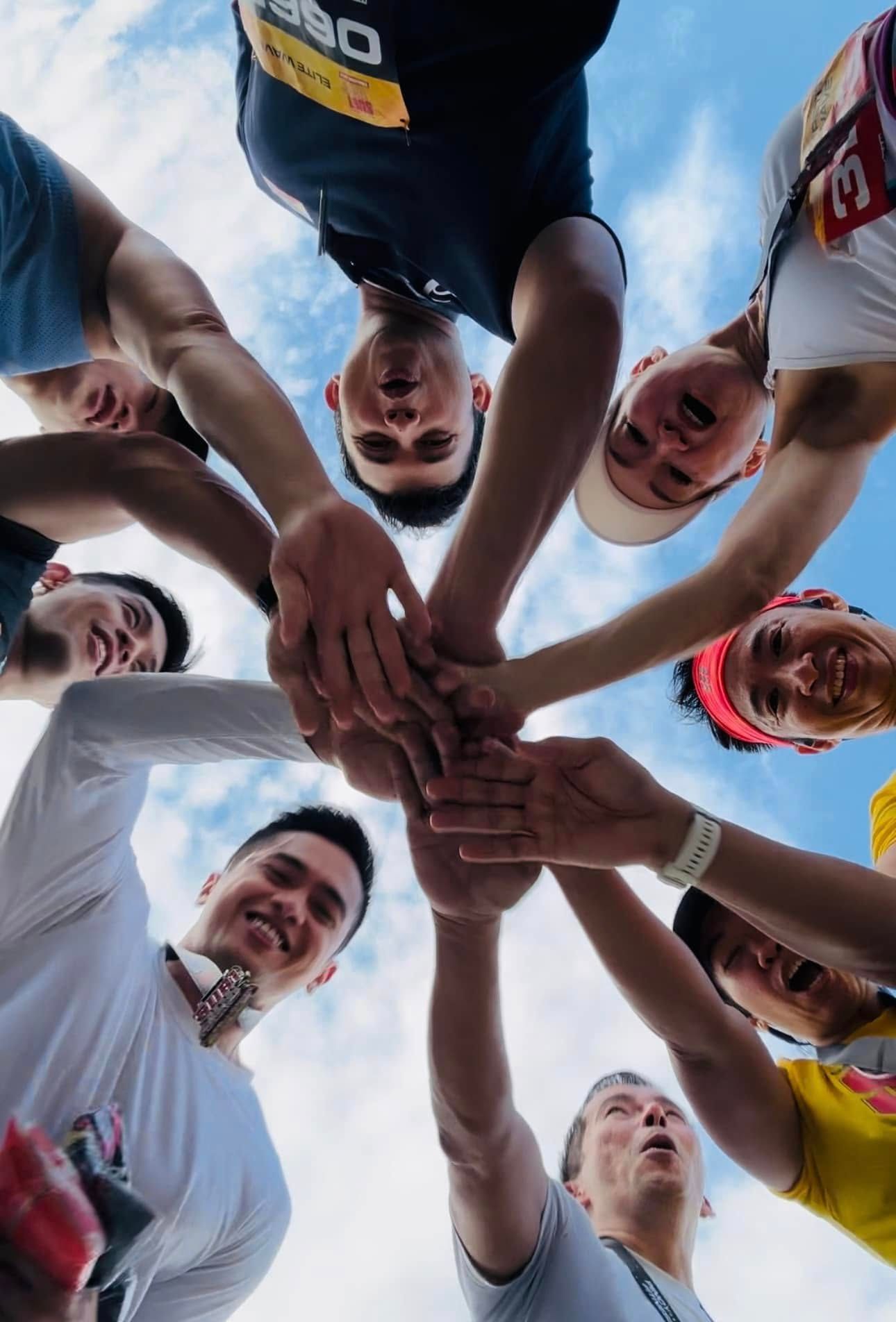 A low-angle view of a group of runners in athletic gear putting their hands together in a circle against a blue sky.