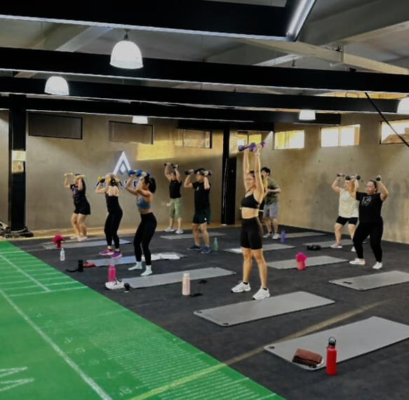 A group of people performing dumbbell overhead presses during a fitness class in a gym with yoga mats on the floor.