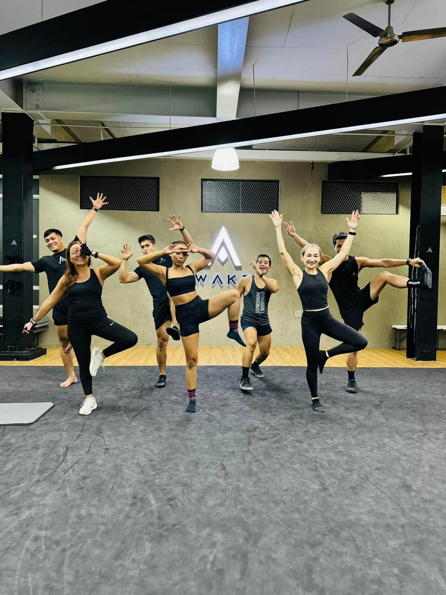 Six people in a gym pose on one leg with arms raised, wearing matching black athletic sets in a studio with an A-logo.