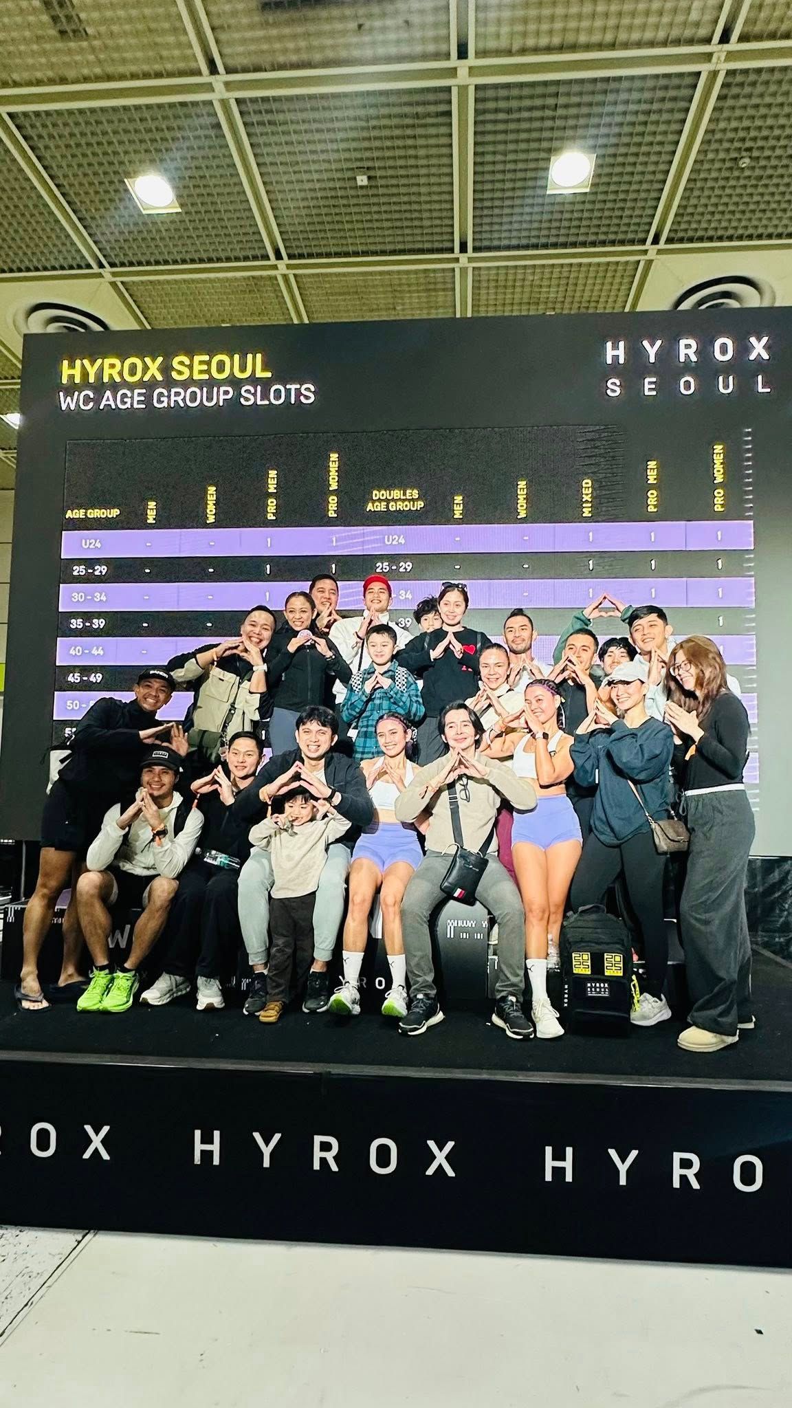 A group of athletes poses on a stage in front of a Hyrox Seoul event banner, smiling and gesturing at the finish.