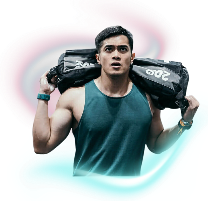 A person in a dark green tank top carrying a black sandbag on their shoulders against a soft, colorful light background.