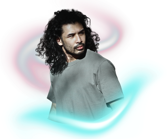 A person with long, dark, curly hair wearing a gray t-shirt, posed against a background of soft, glowing light swirls.