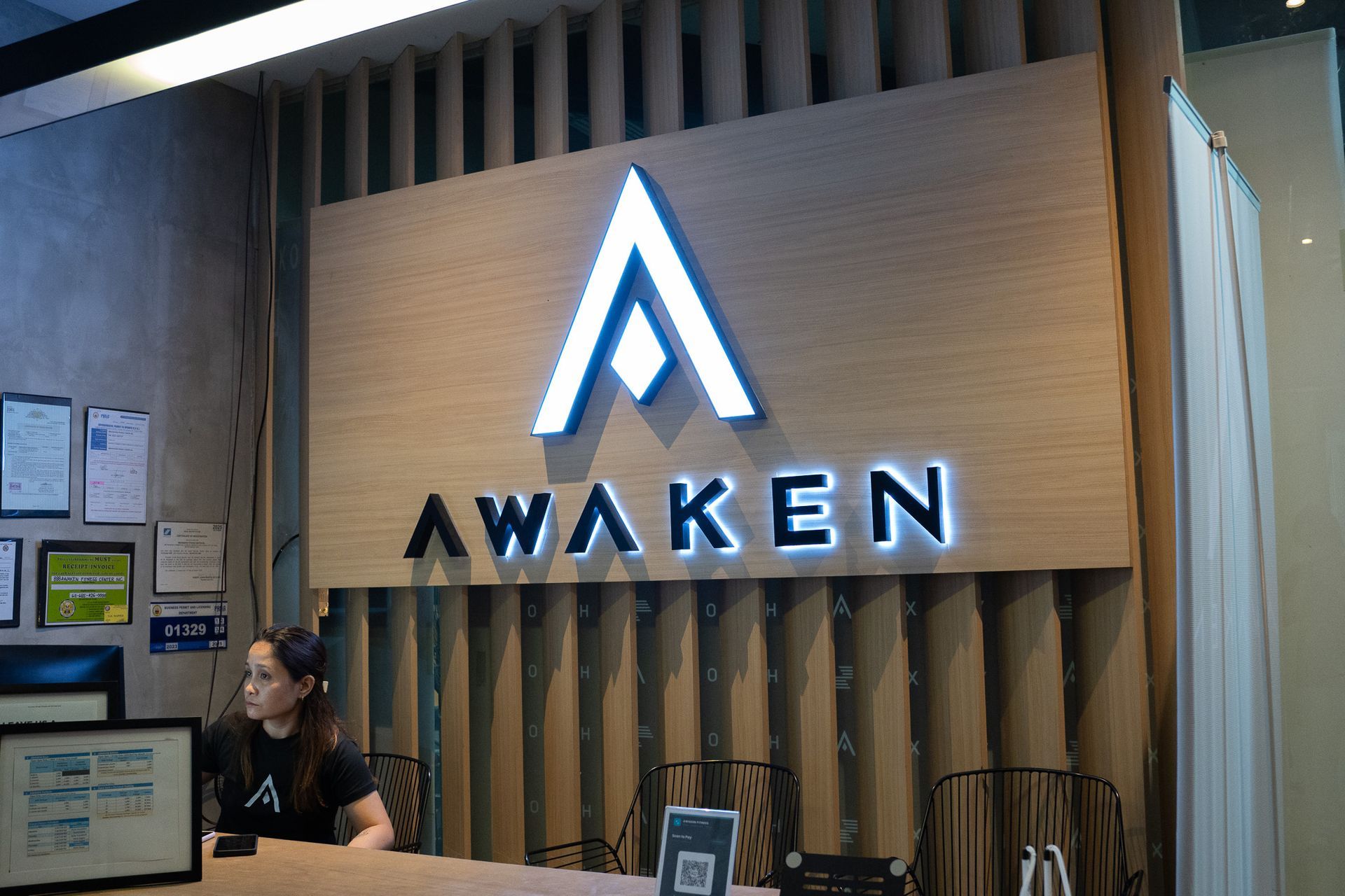 Reception desk with illuminated AWAKEN logo on a wood wall, and a staff member seated at left.