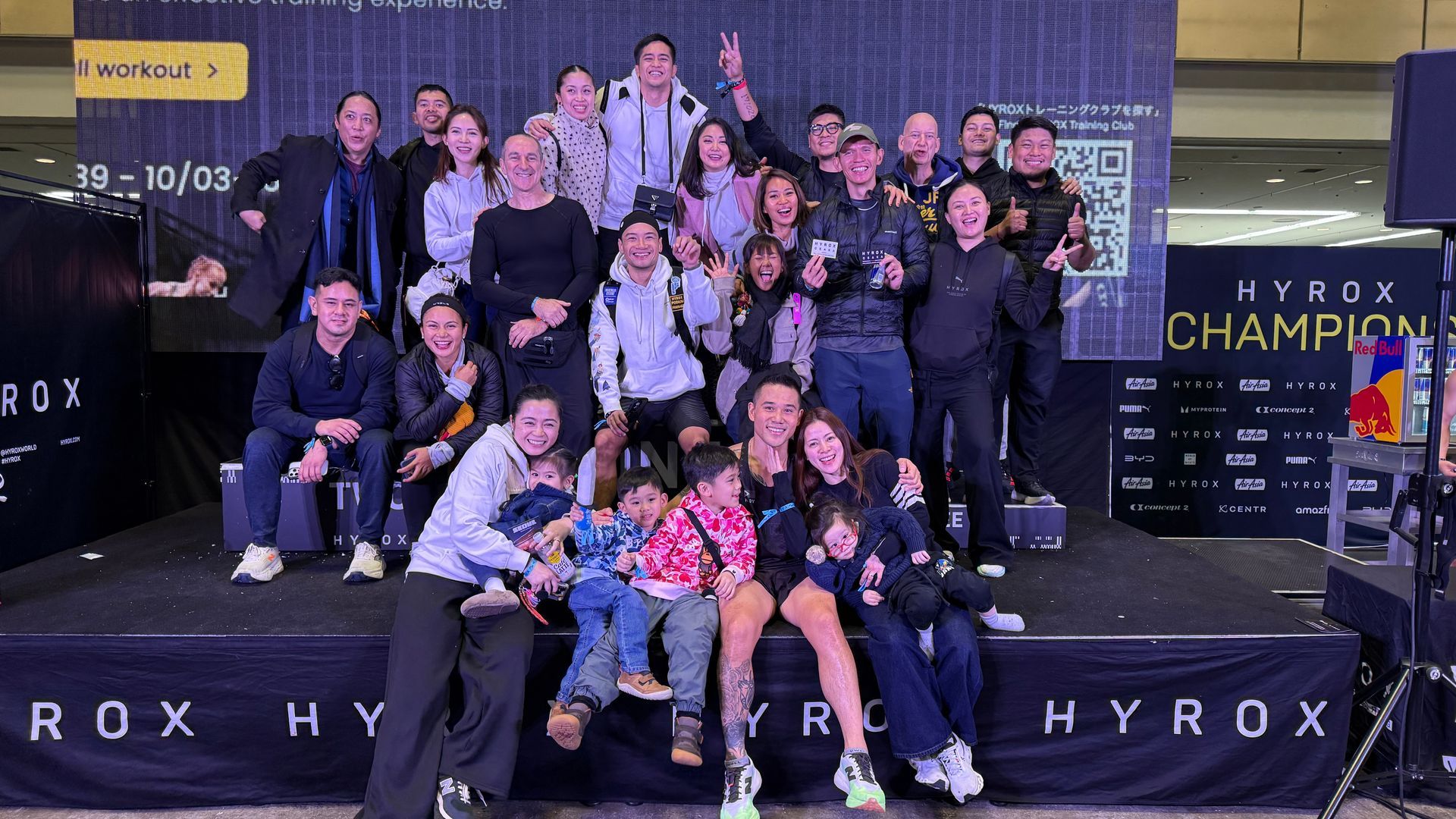 A large group of people posing for a photo on a stage at a Hyrox competition with black banners in the background.