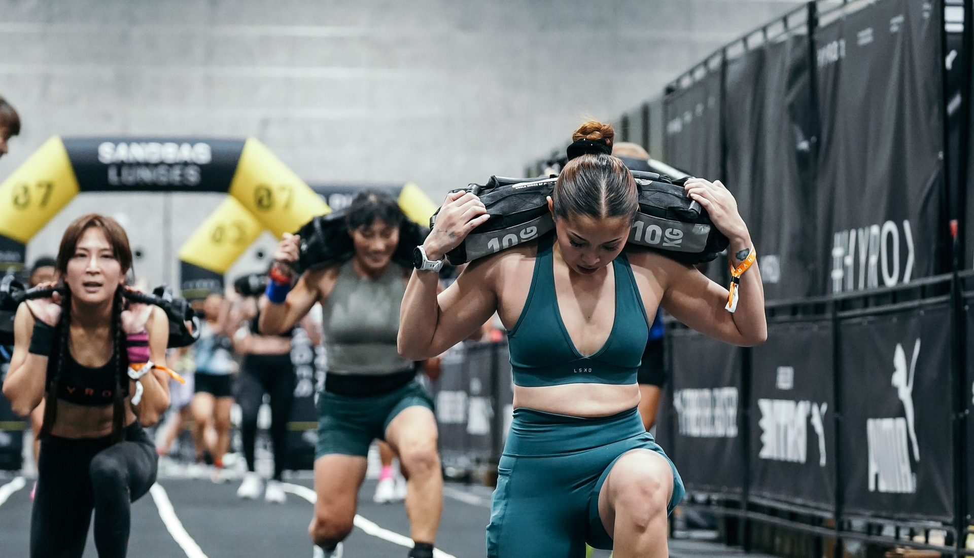 Athletes in activewear perform weighted sandbag lunges at a fitness competition under a yellow archway.