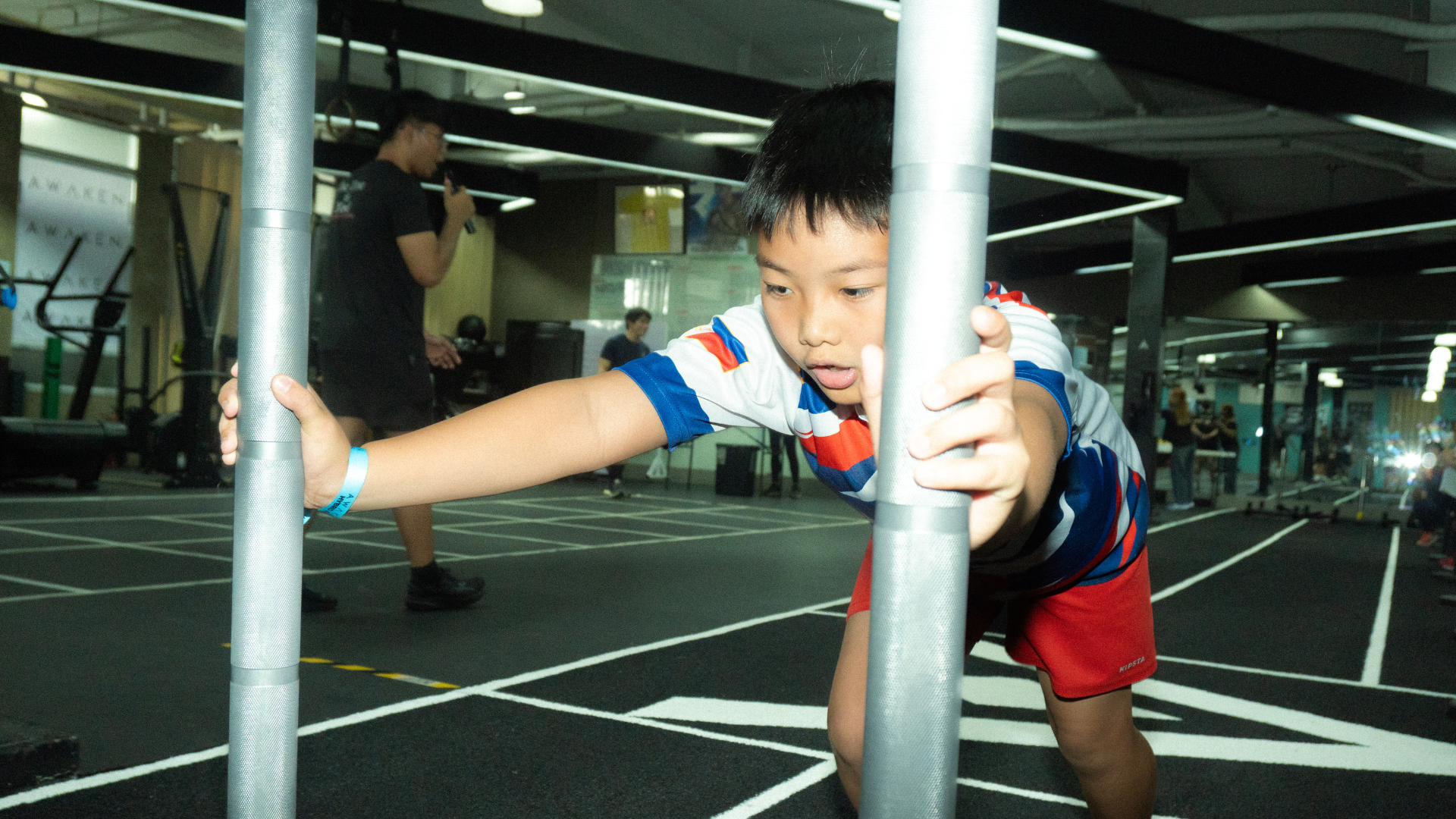 Child stretching between metal bars in an indoor gym, with another person in the background.