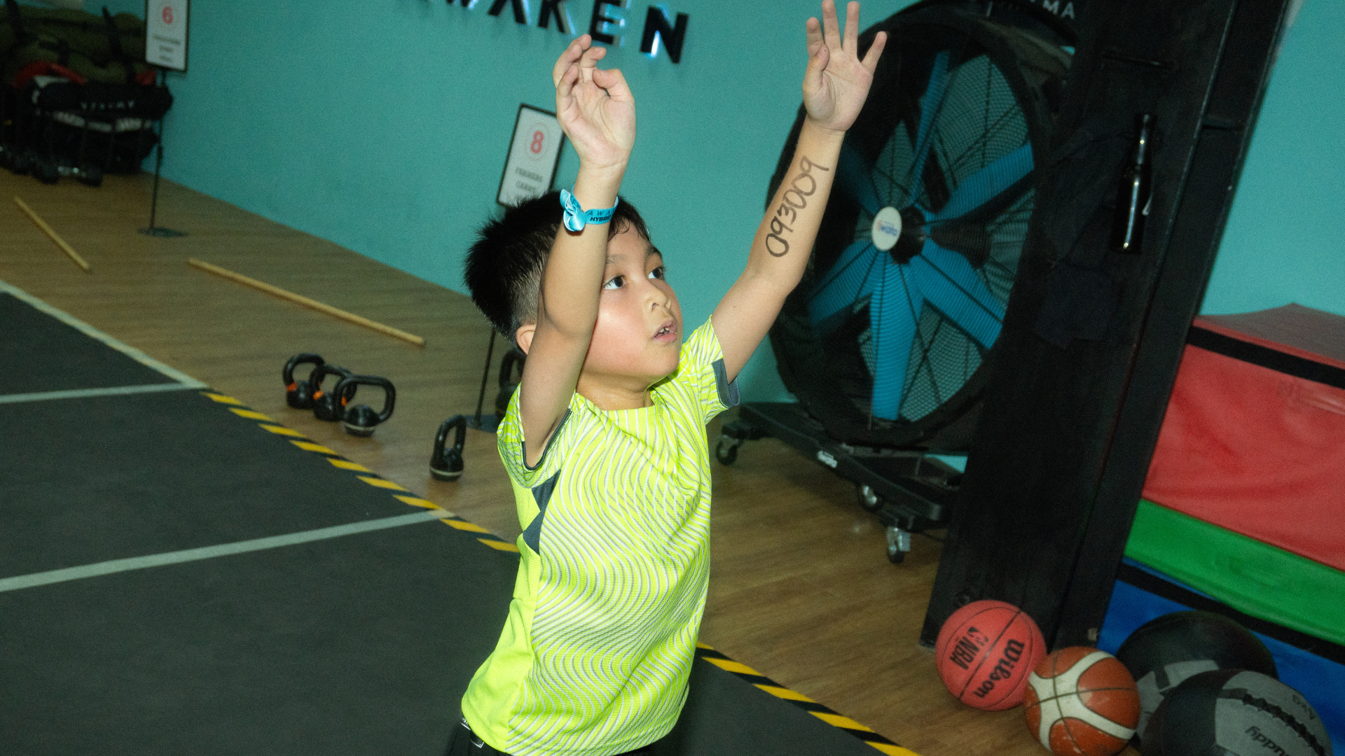 Child in neon shirt reaching both arms up in a gym, with exercise equipment in the background
