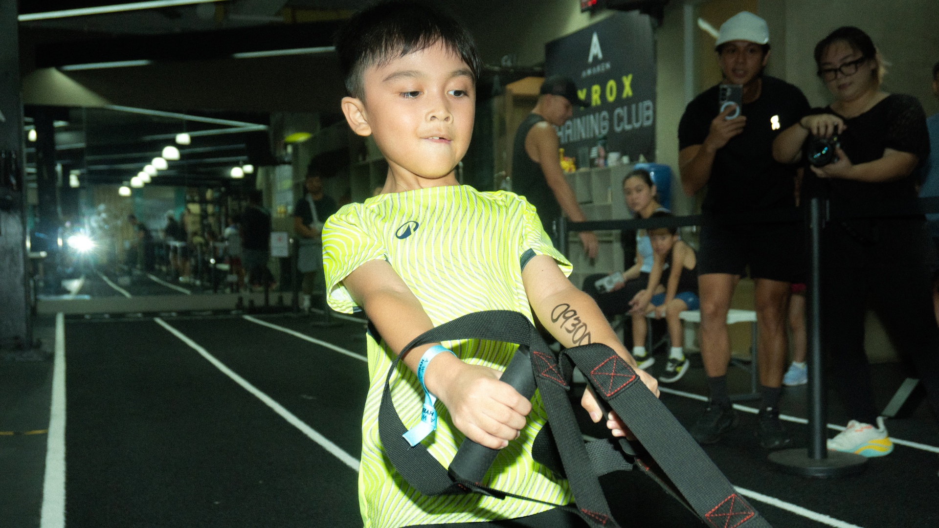 Child in neon yellow shirt pulling black straps in an indoor event space, with spectators in the background