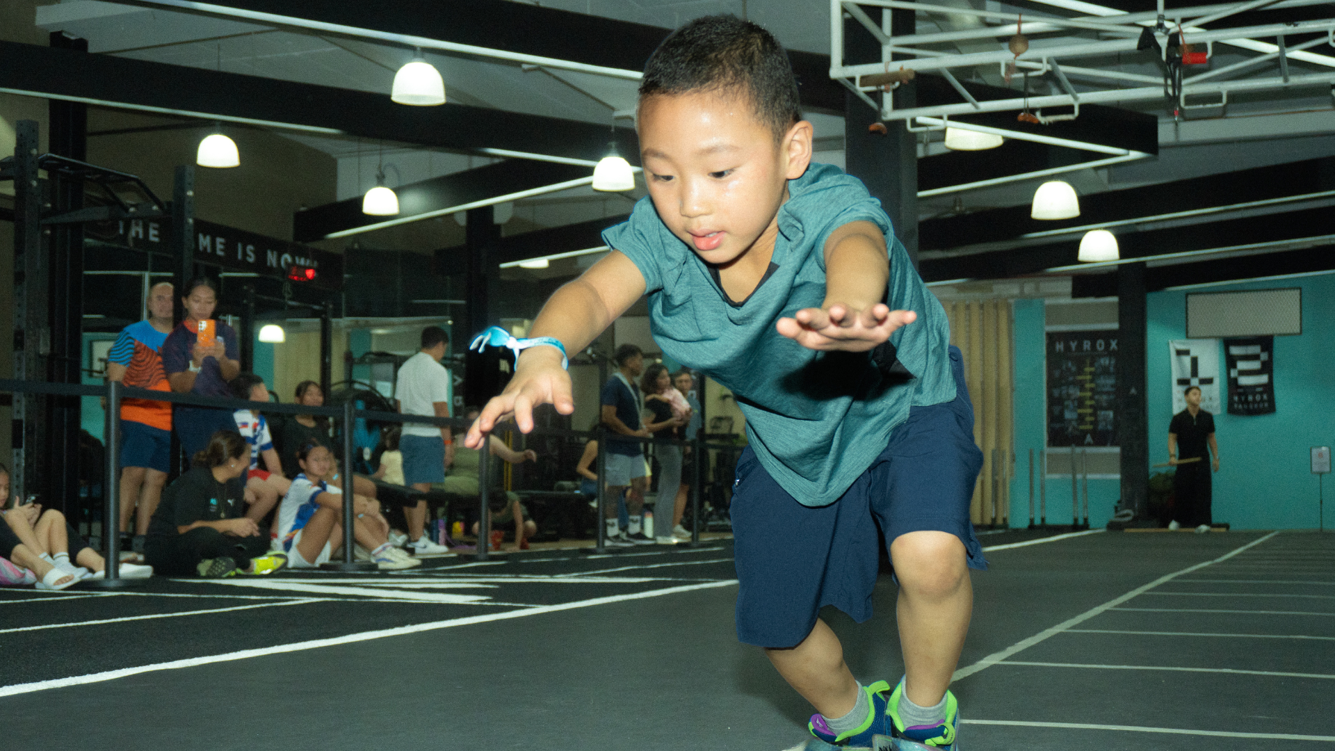 Person sprinting indoors on a gym track with arms outstretched, wearing a teal shirt and navy shorts.