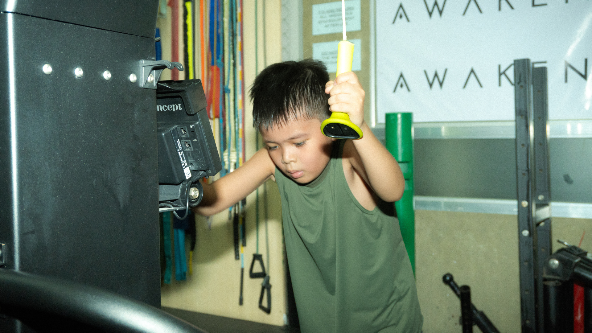 Person in green shirt leaning on exercise equipment in a gym, holding a yellow object near their head.