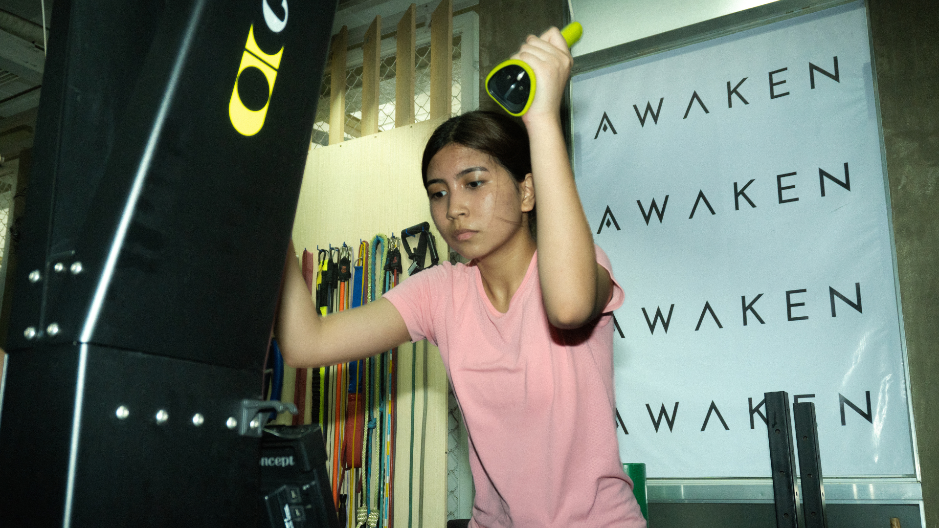 Person in pink shirt exercising with arms raised in a gym studio with “AWAKEN” banners.