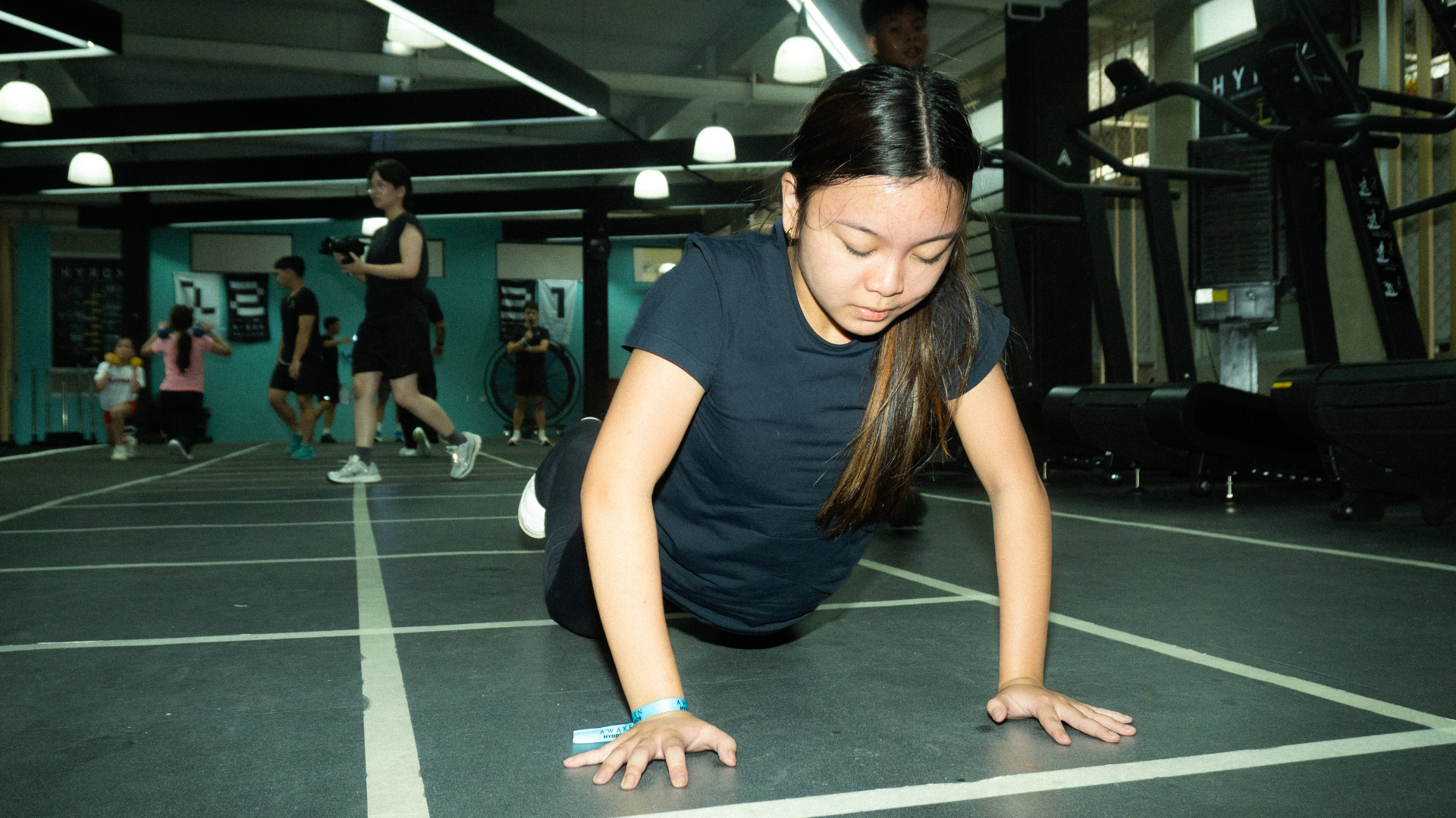 Person doing a push-up in a gym, focused on the floor with other people working out in the background.