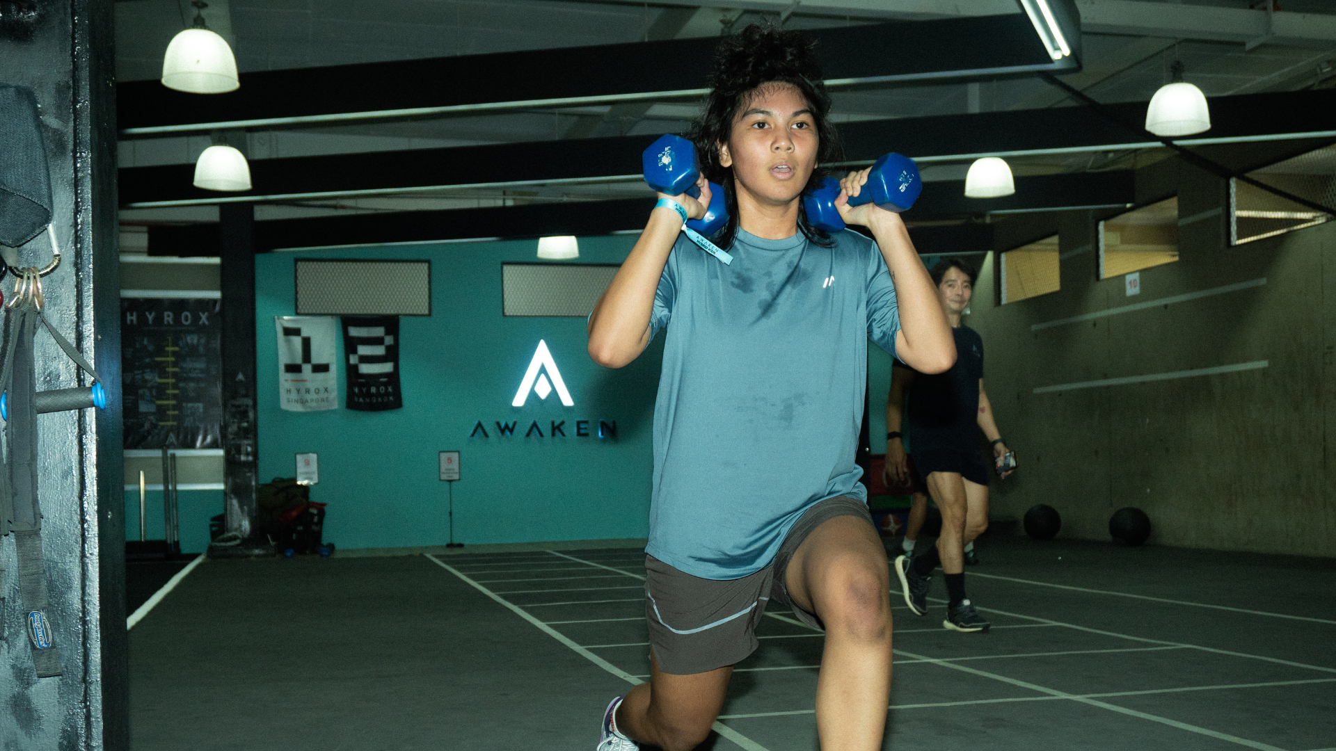 Person exercising indoors, holding blue dumbbells while lunging on a gym floor.