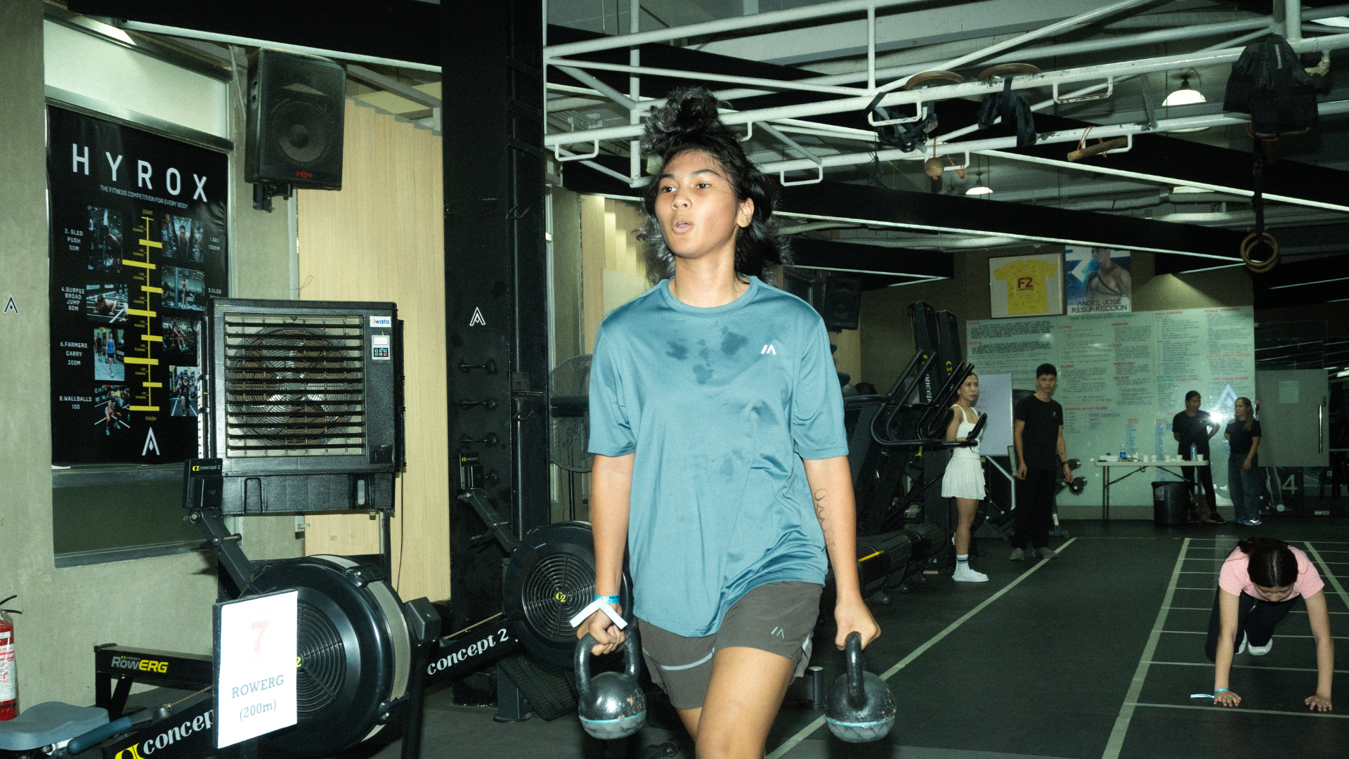 Person walking in a gym carrying a kettlebell, with exercise equipment and mirrors in the background.
