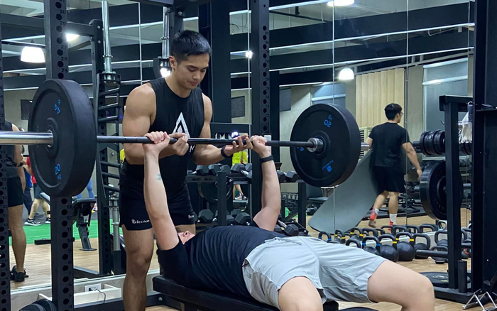 A spotter stands behind a person performing a barbell bench press inside a gym.