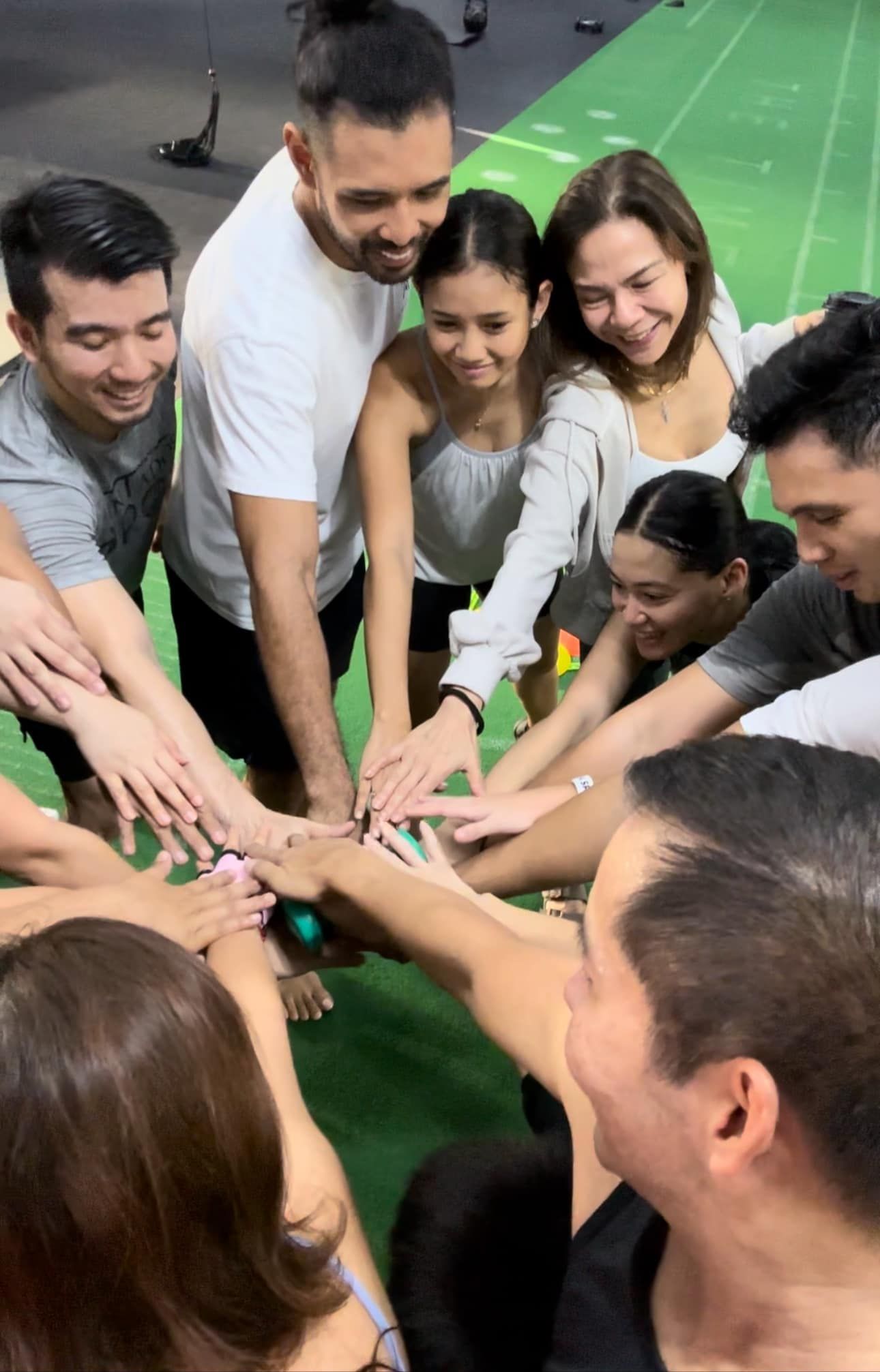 A group of people gathers in a gym, smiling and placing their hands together in the center to show teamwork and unity.