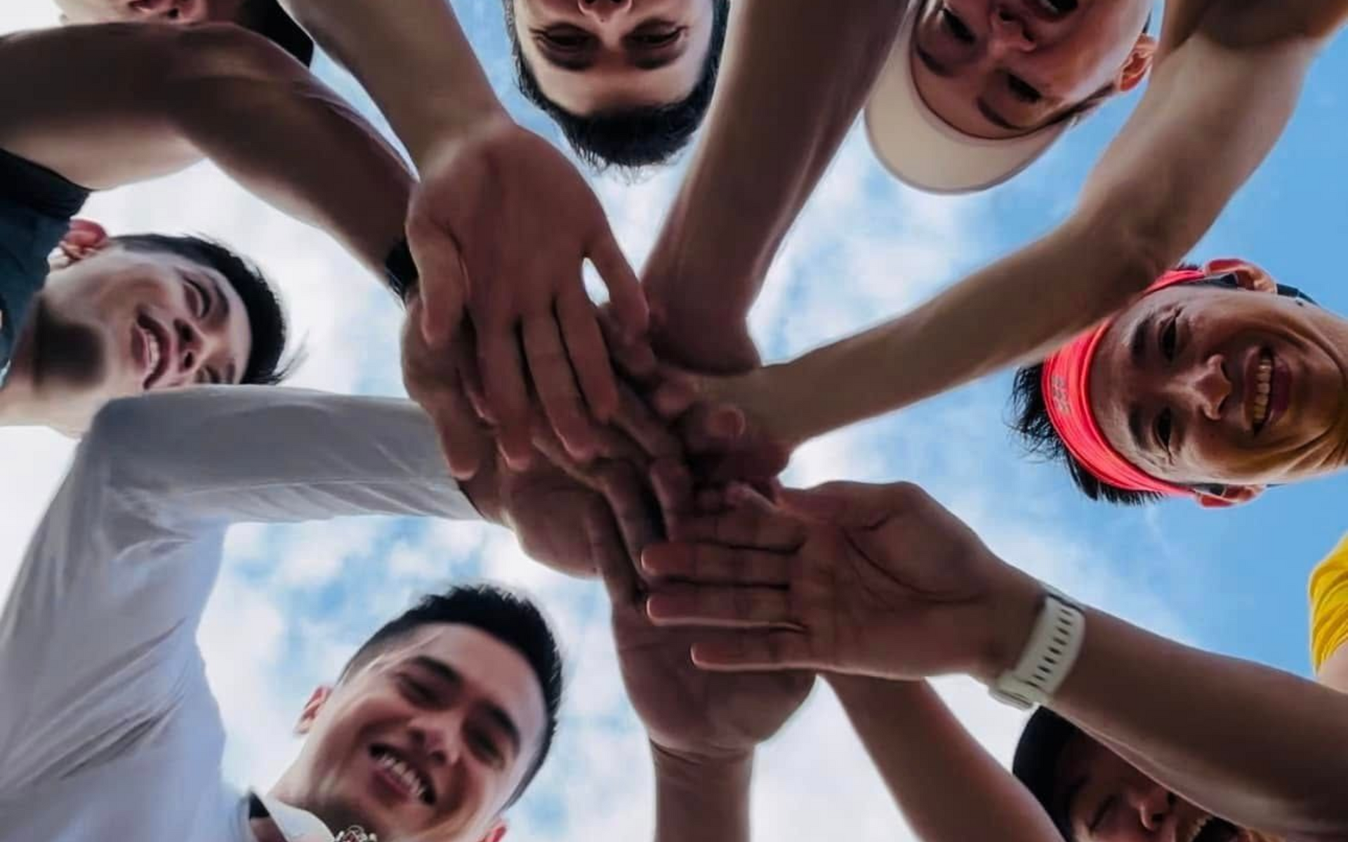 A group of people look down at the camera with joyful expressions, their hands stacked together against a blue sky.