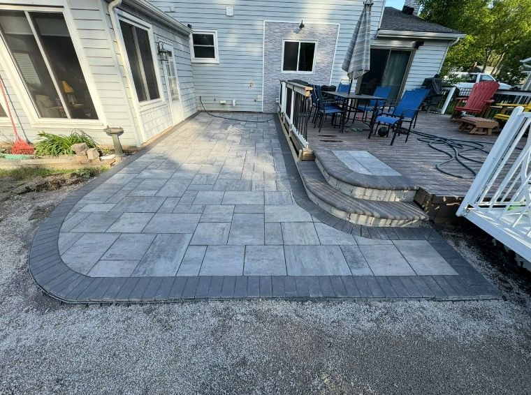 Newly installed gray paver patio with matching curved border, leading to a deck with outdoor furniture.