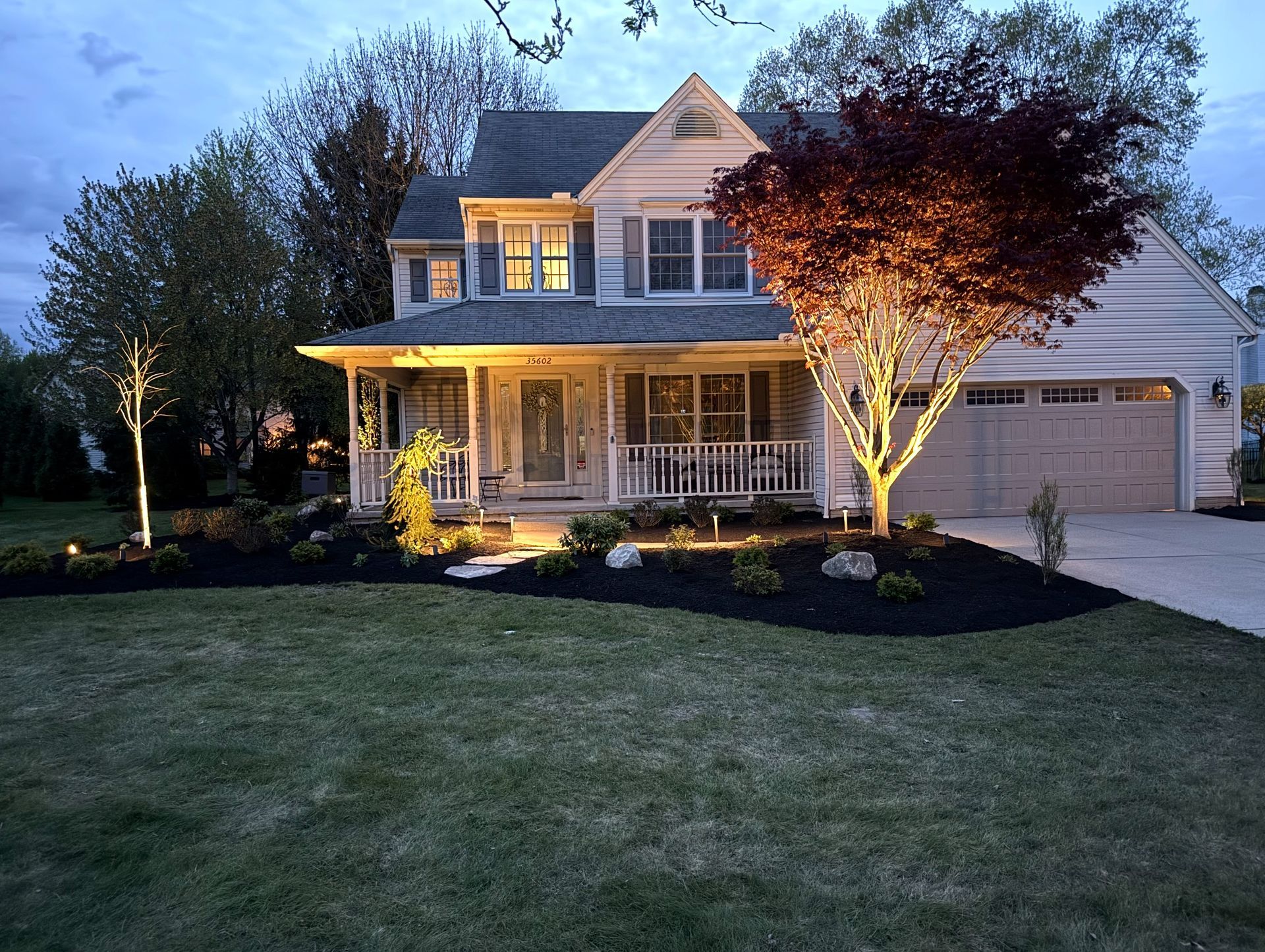 Two-story house at dusk, lit by exterior lights, with a porch and landscaped yard.