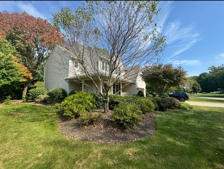 Two-story house with beige siding, surrounded by green shrubs, trees, and grass, under a blue sky.