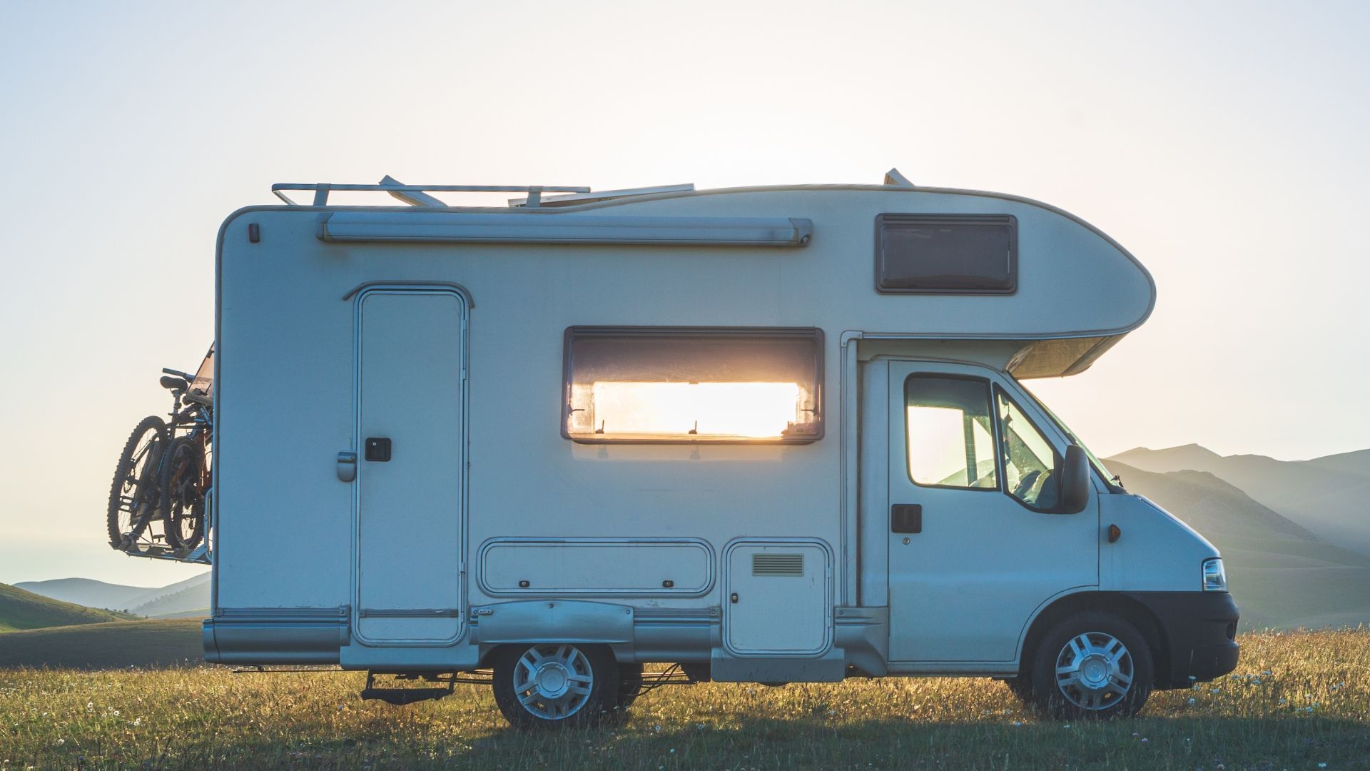 White RV parked in grassy field with sun setting behind it. A bicycle is mounted on the back.