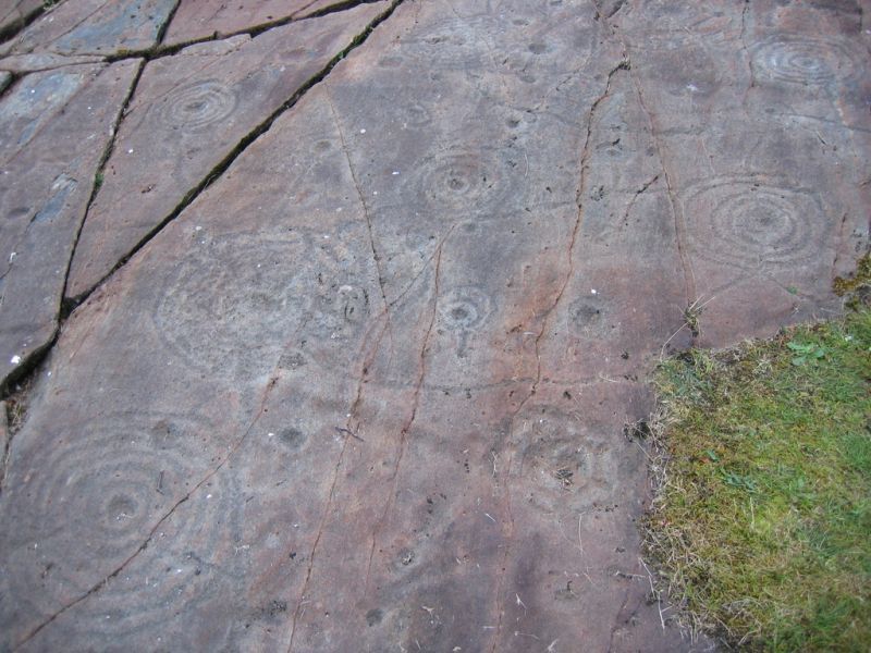 Petroglyphs carved into a rock surface, featuring concentric circles and lines. Brown stone with some grass.