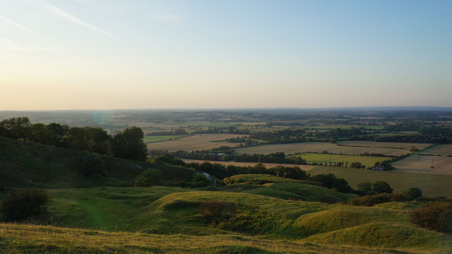 Rolling green hills overlooking a vast farmland landscape under a pale blue sky.