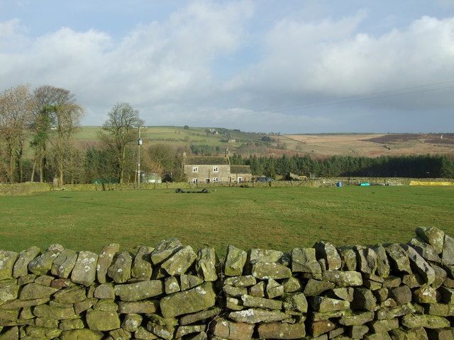 Green field with a stone wall in the foreground, a house, and hills under a cloudy sky.