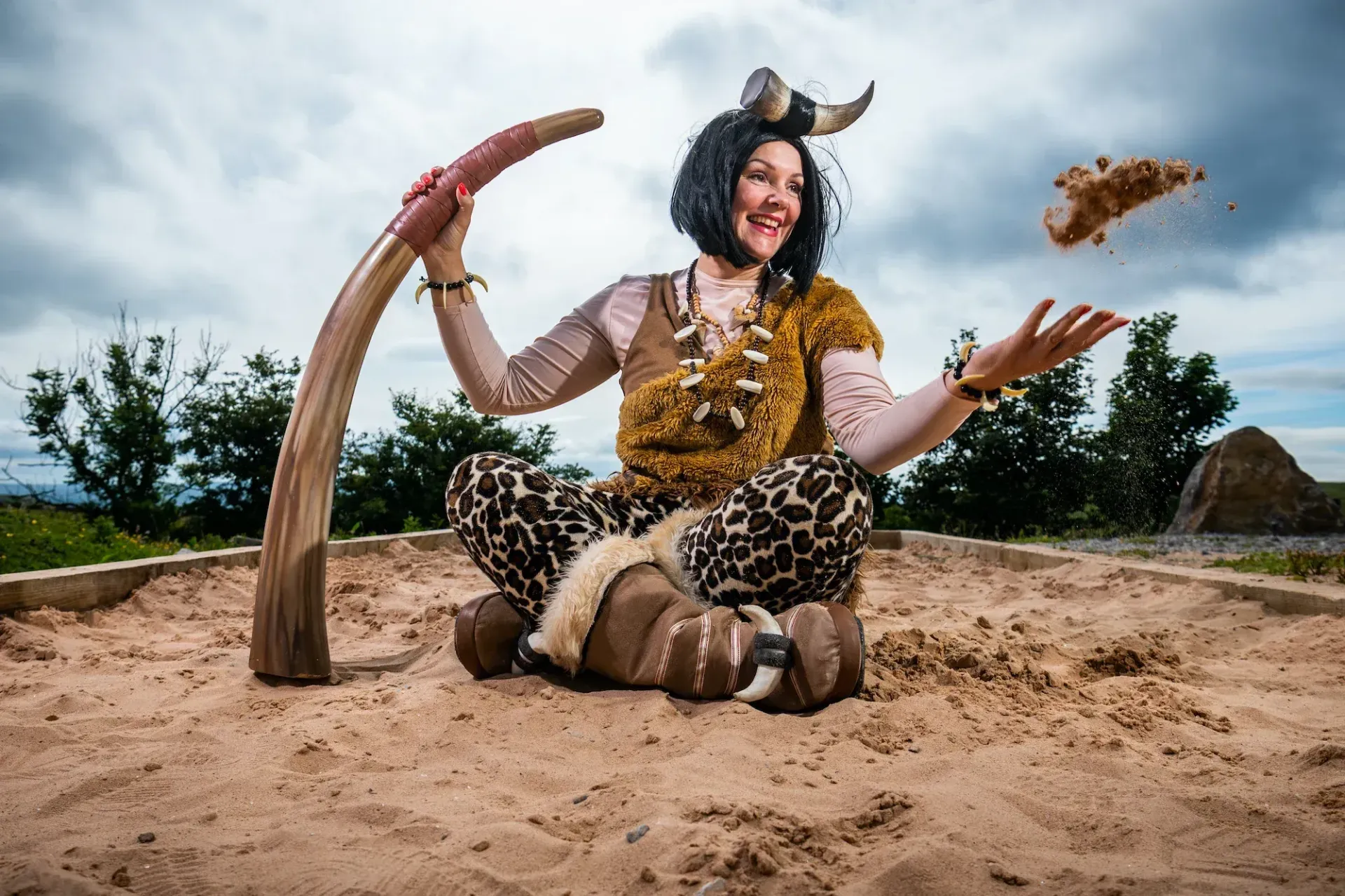 Woman in cavewoman costume sitting in a sandbox, tossing sand in air, holding a wooden club.