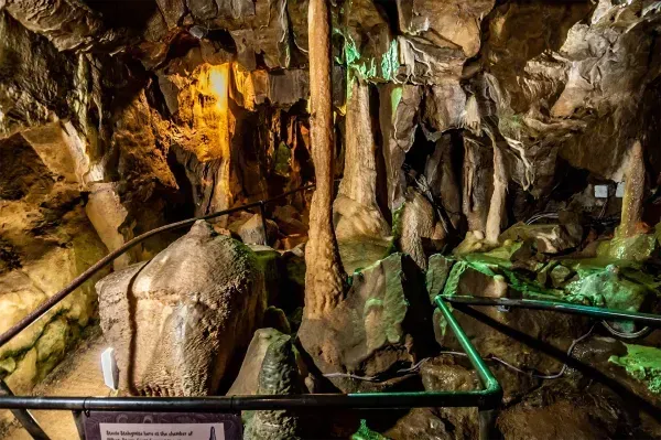 Inside a cave, stalactites and stalagmites are illuminated by yellow and green lights. Black railings guide visitors.