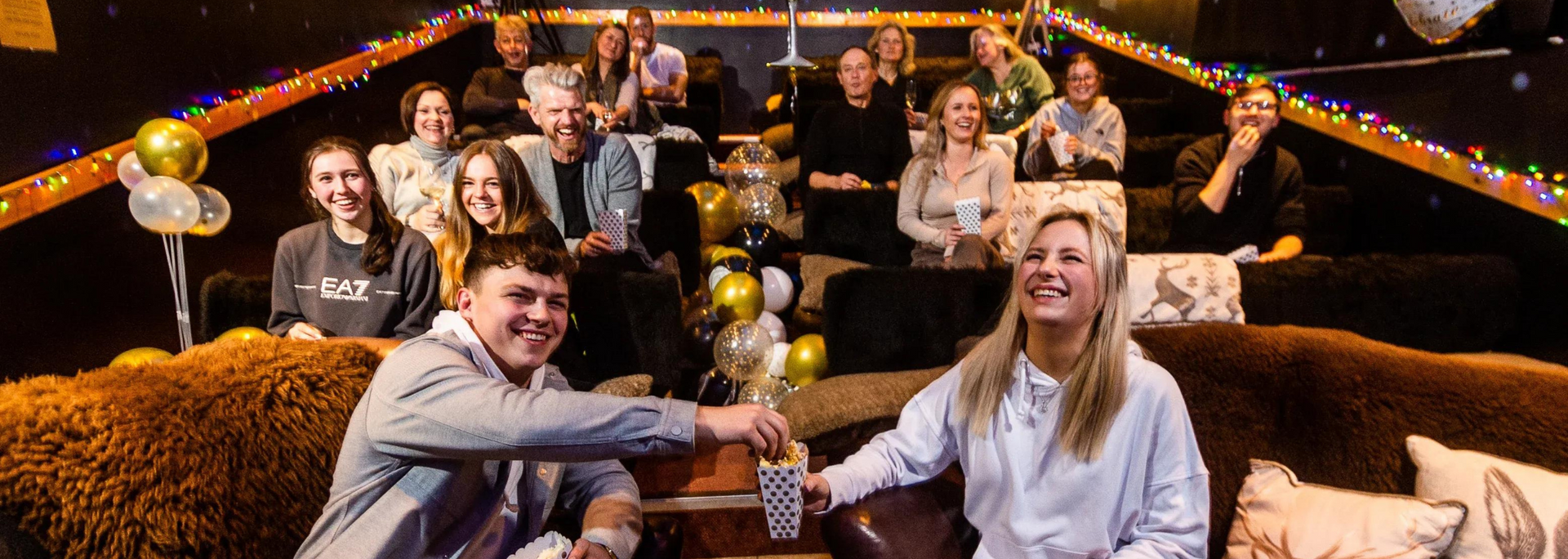People laughing in an auditorium, a man and woman holding cards, balloons present.