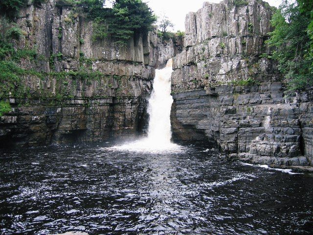 Waterfall cascading into a dark pool, surrounded by layered gray rock cliffs, in a natural setting.