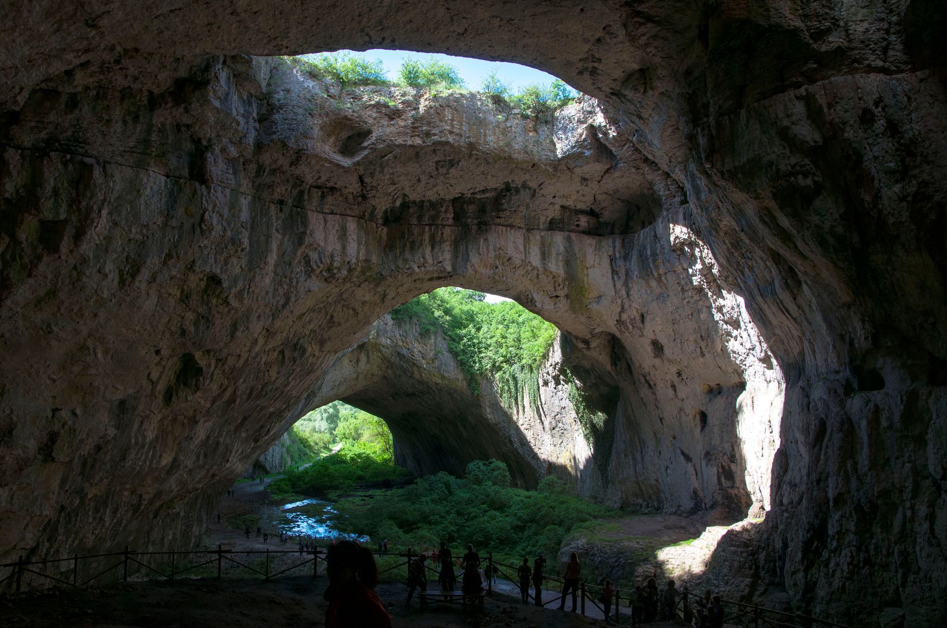 Large cave interior with natural arches, sunlight streams in, people visible.