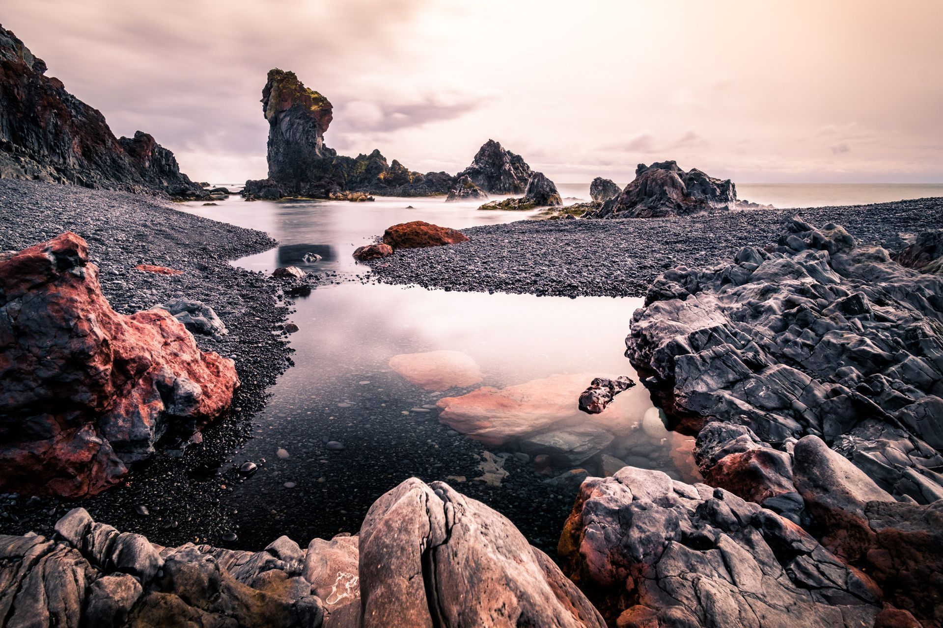 Rocky coastal landscape with black sand, dark rocks, and a cloudy sky reflecting in the water.