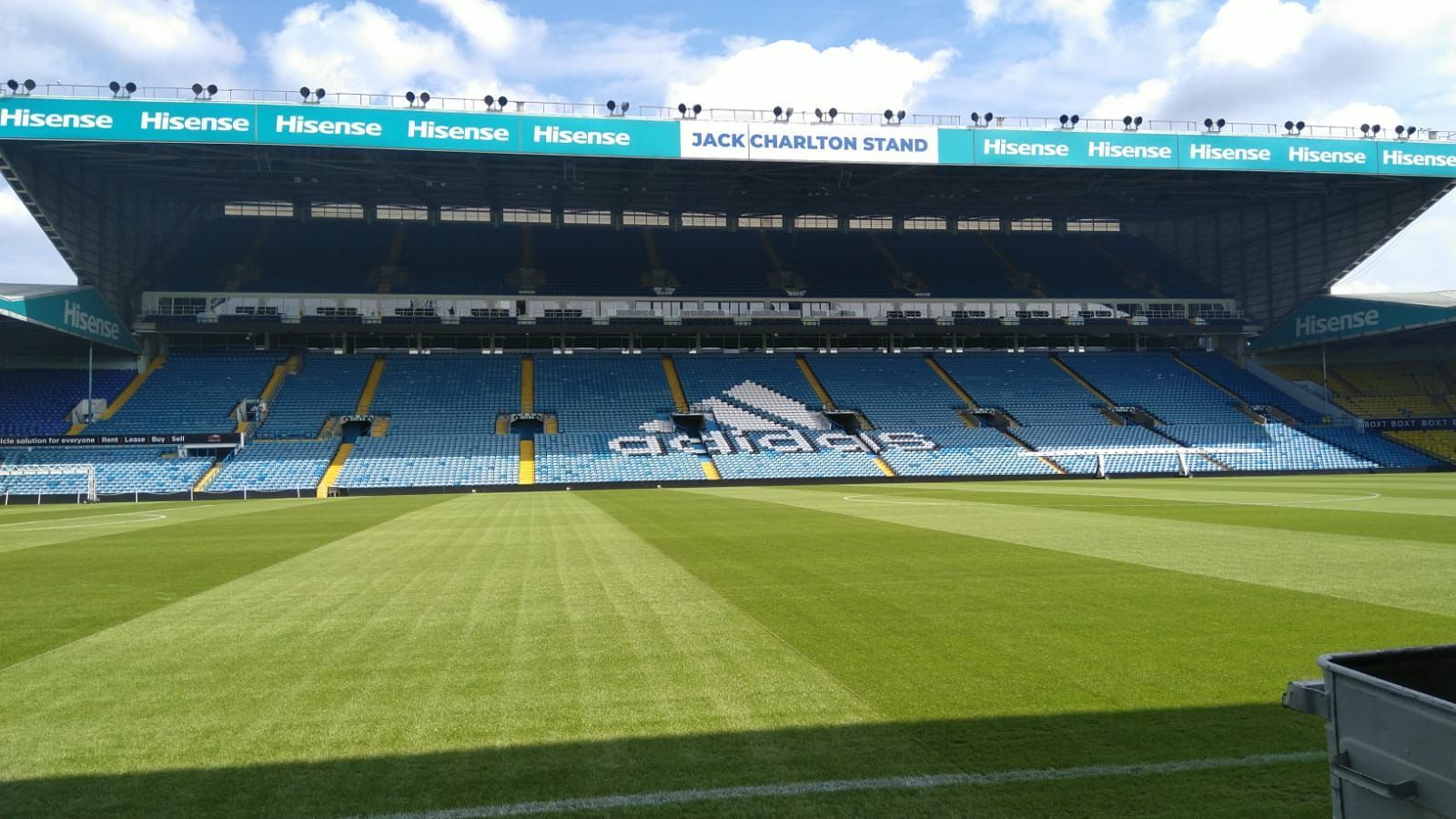 Empty blue stadium seating, green pitch, clear sky. 