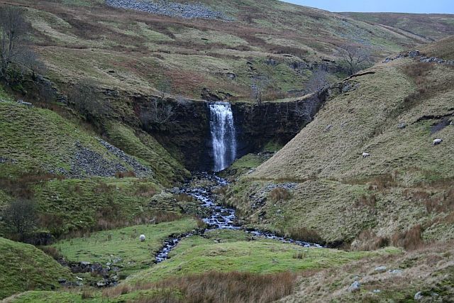 Waterfall cascading down a rocky cliff into a stream, surrounded by grassy hills.