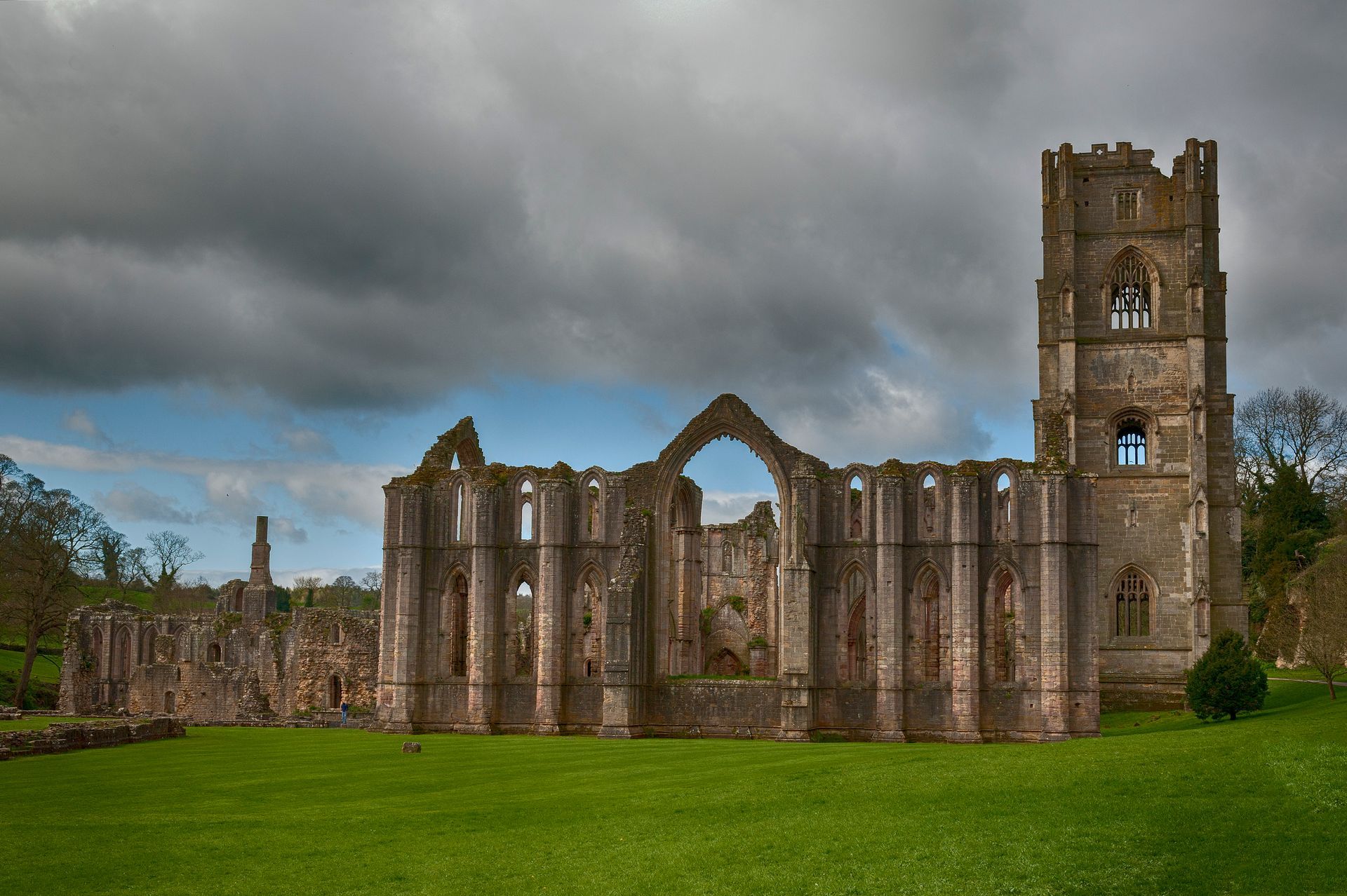 Ruined stone abbey with a tall tower under a cloudy sky. Green grass in the foreground.
