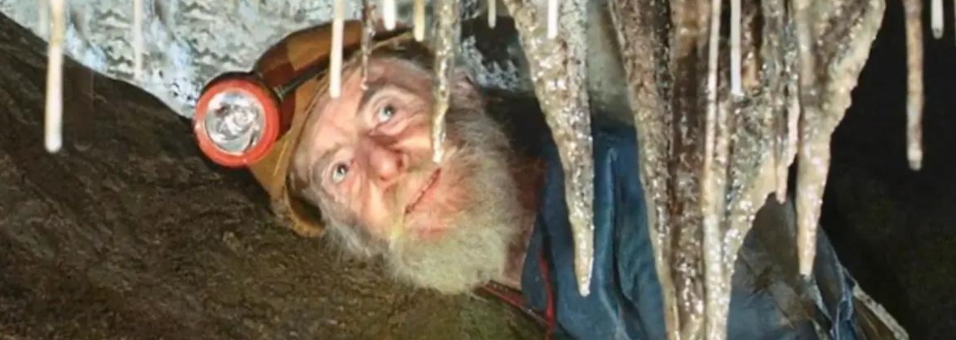 Man with headlamp explores a cave, looking up at stalactites.