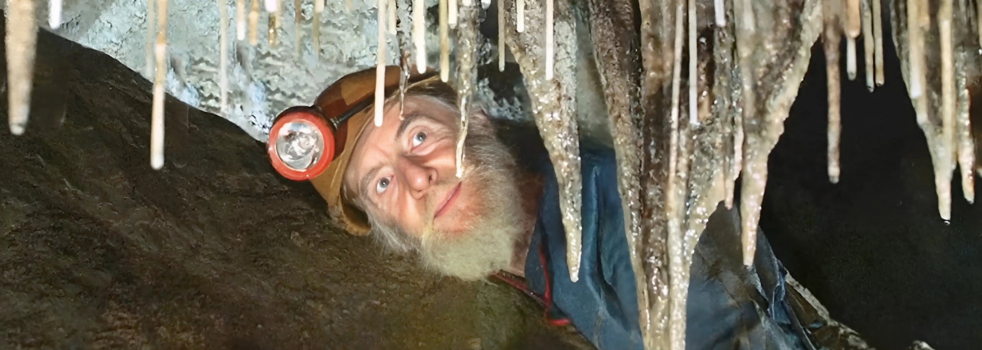 Man in cave with headlamp, looking up at stalactites.