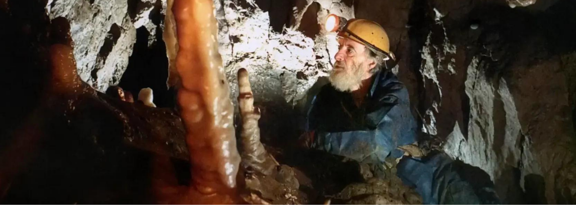 A person in a cave, wearing a helmet, examining a stalagmite.