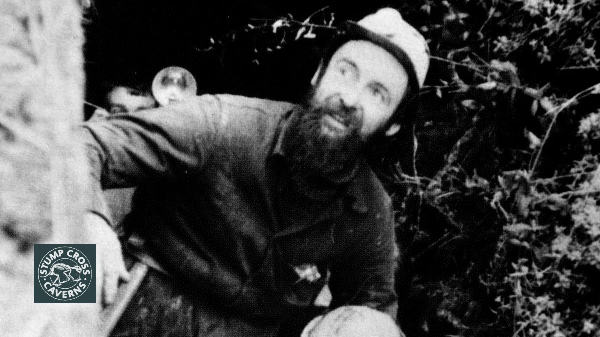 Man with beard and hardhat emerges from dark space, clutching something, with foliage around.
