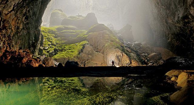 Large cave with person silhouetted, reflected in still water, sunlight streaming through opening.